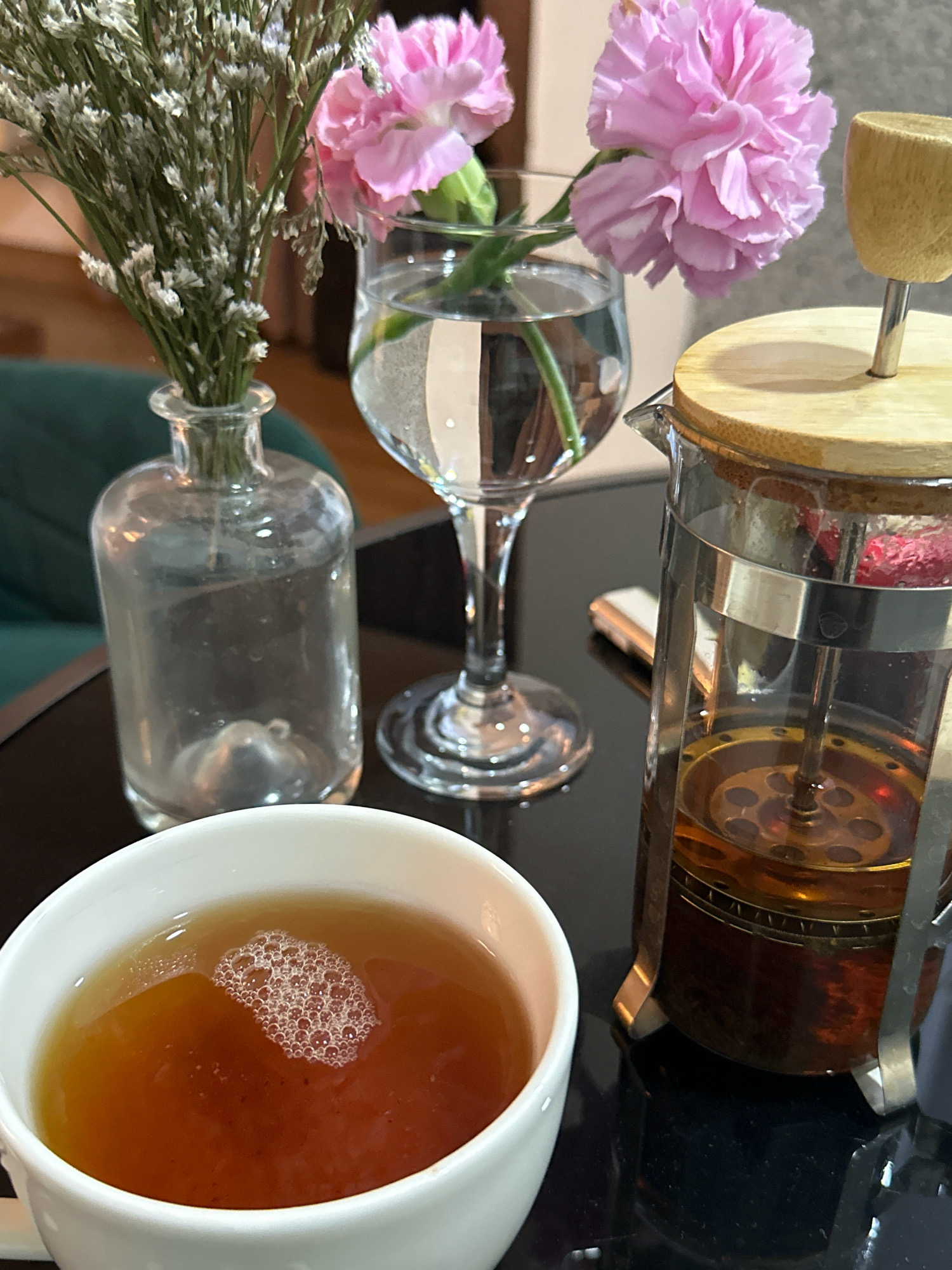 A high-angle close-up shot captures a serene setting with a cup of tea, a French press, and a small floral arrangement. The cup, filled with a light amber liquid, is in the foreground, its surface dotted with small bubbles. To its right sits a glass French press containing a darker amber liquid and hints of dried fruit, topped with a light wooden lid. Behind these, a delicate arrangement of dried white flowers in a clear glass bottle is visible, juxtaposed with two vibrant pink carnations in a stemmed glass of water. The scene is set on a dark, reflective surface, suggesting a table. The background is softly blurred, showing hints of a green upholstered chair and a wooden floor, implying an indoor environment, possibly a cafe or a home. The image has a soft, natural light that highlights the textures and colors of the subjects. There are no people in the image, and the overall atmosphere is peaceful and inviting. The image appears to be taken in Lalitpur, Nepal, given the context of the prompt.