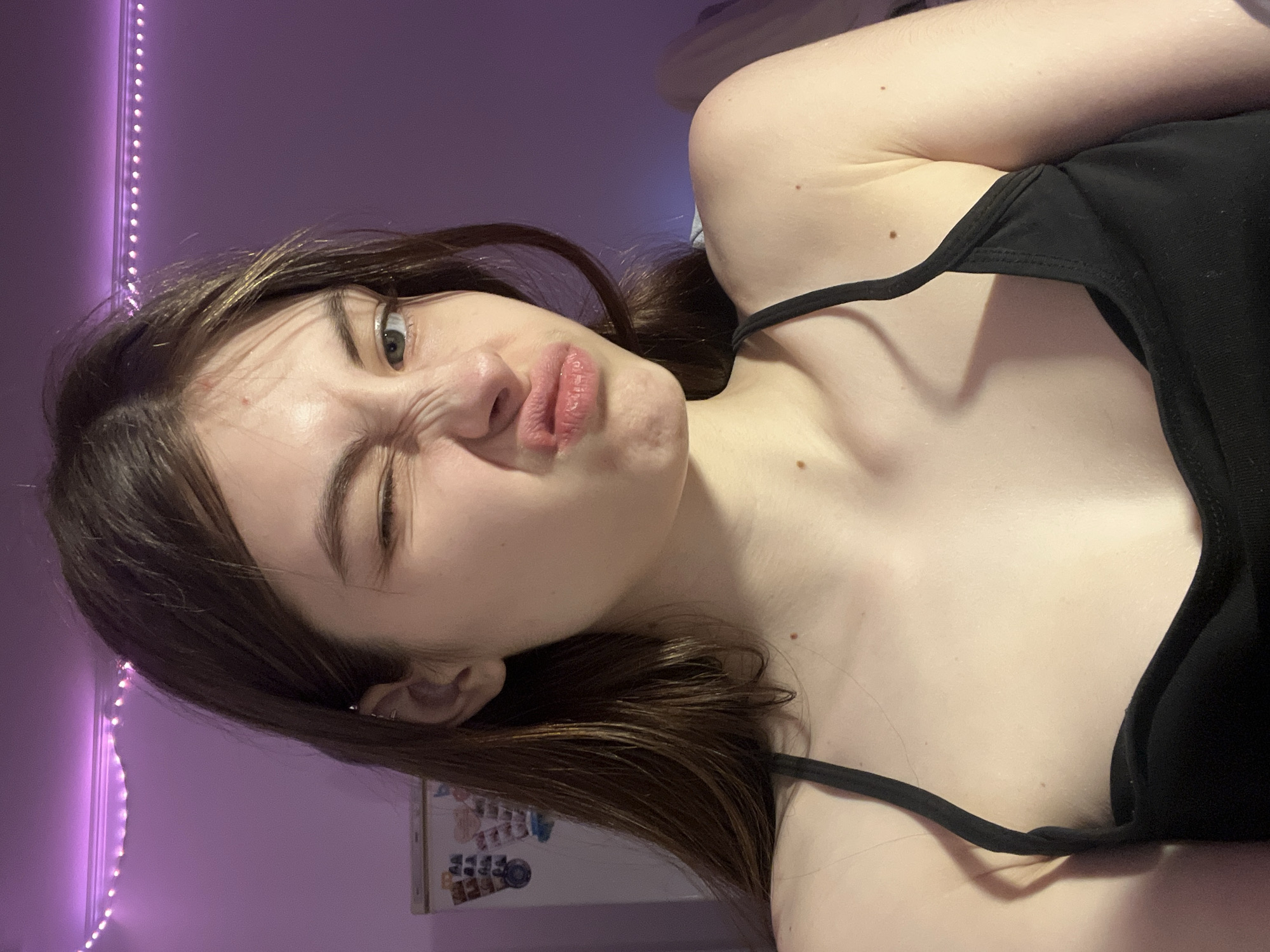 A young woman with brown hair and fair skin is posing for a selfie. She is wearing a black tank top. Her face is contorted into a playful grimace, with one eye squinted shut and her lips pursed. The background is a blurred purple wall with a string of LED lights along the edge. A refrigerator or bulletin board with various papers and items attached is visible in the lower background. The location cannot be determined from the image, and it is not possible to ascertain whether the photo was taken in Saint Petersburg, Russia.