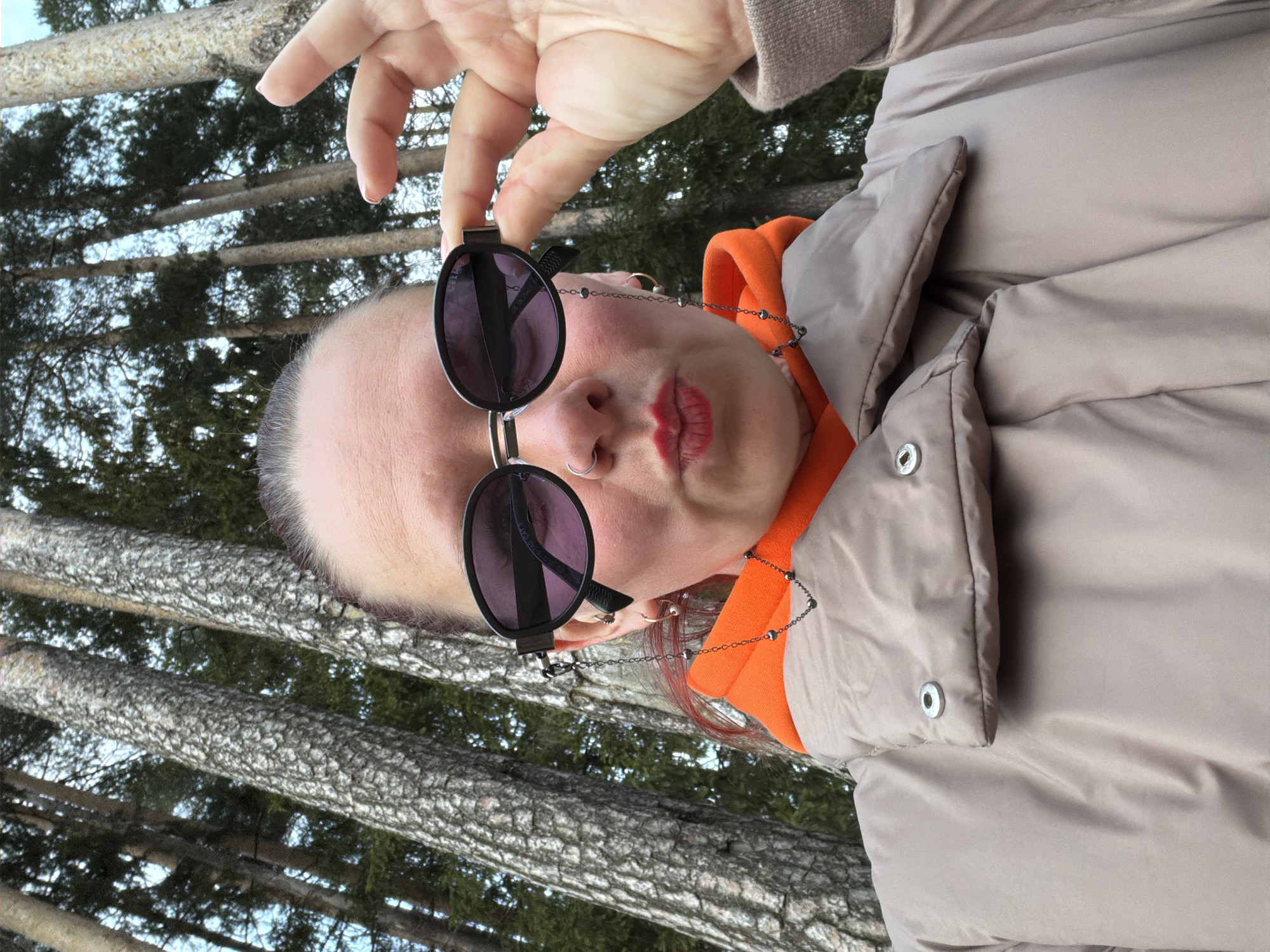 A person is holding sunglasses in their right hand, with trees in the background. The person is wearing a beige puffer jacket over an orange hooded sweatshirt. The person has a nose ring and multiple earrings. The background appears to be a forest with tall, bare trees. The location may be Chaikovsky, Russia based on the prompt.