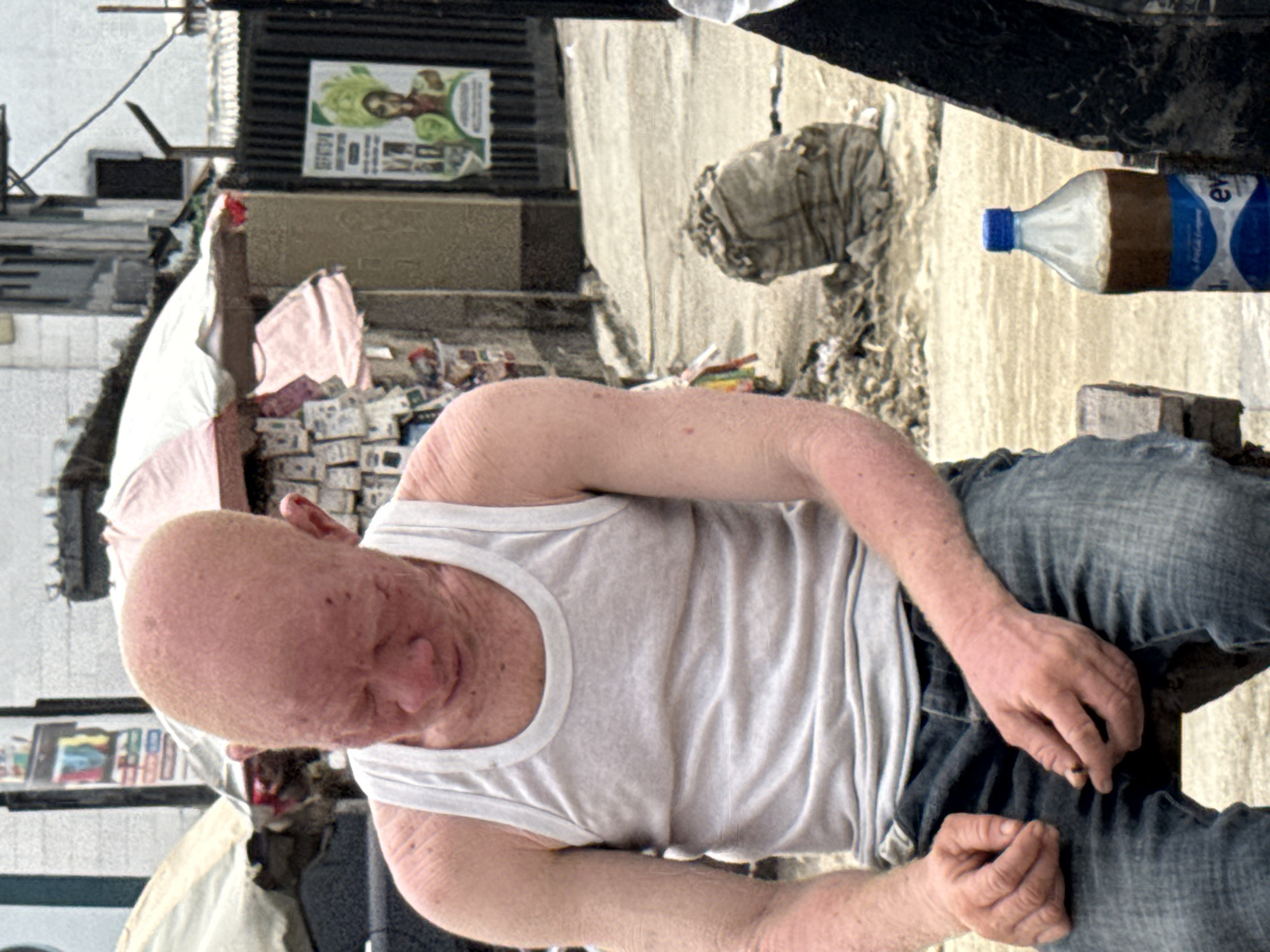 The image, when correctly oriented, features an individual with albinism seated outdoors in what appears to be a street or market setting in Ado, Nigeria. The person, viewed from slightly above, is wearing a white tank top and blue jeans, looking downwards with a calm or contemplative expression. Their skin is notably pale, consistent with albinism, and their hair appears very light. Their hands rest in their lap, with one hand holding a small, dark object.
In the mid-ground, a plastic bottle with a blue cap, containing a brown liquid, is visible on the ground. The label on the bottle shows partial text including "EV" and "EVE", likely referring to a brand like Eva, along with a blue logo and "PET."
The background reveals an active outdoor environment under bright daylight. Behind the seated person, stacks of small items, possibly bundles of goods or currency, suggest a vendor's stall or a commercial area. Further back, buildings with visible details, including a poster on a wall, frame the scene. The poster depicts a person surrounded by green leaves, with some unreadable text below. The ground surface is uneven, a mix of dirt and worn pavement, consistent with an urban or semi-urban street. The overall impression is one of daily life in a bustling outdoor location.