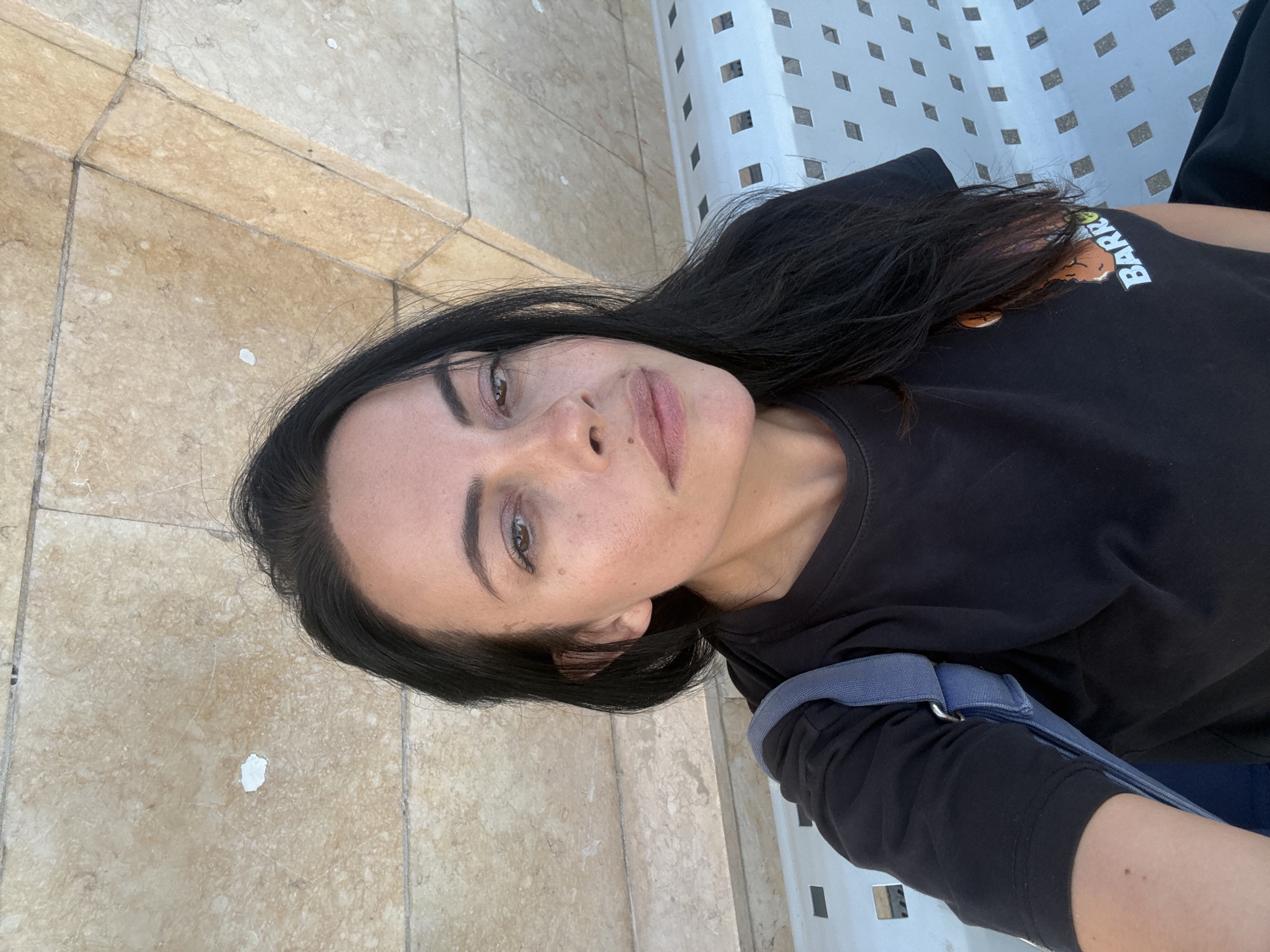 A medium shot captures a woman with dark hair, a dark t-shirt, and a blue bag strap across her shoulder, sitting on a white bench. The background consists of a light-colored tiled wall. The bench has a diamond-shaped pattern. The lighting suggests an outdoor setting during the day.