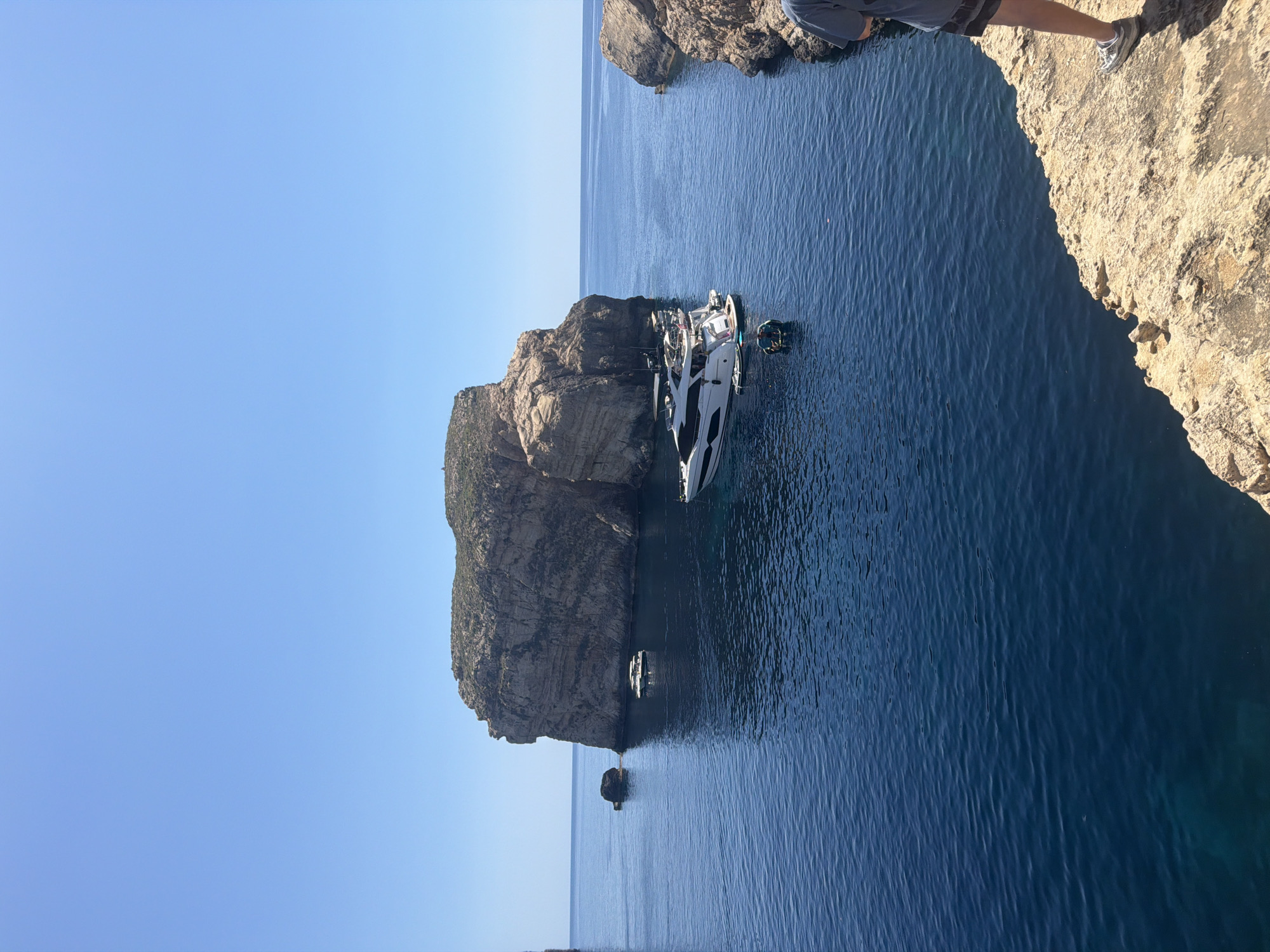 The image depicts a bright, sunny day with a clear blue sky over a coastal area. In the foreground, a person's legs and feet, wearing shorts and sneakers, are visible on a rocky, light-colored cliff edge. The cliff overlooks a body of deep blue water.
In the water, several boats are present. A large white yacht with multiple levels and a dark roof is anchored. Near the yacht, a smaller inflatable boat with people in it is visible. Further out in the water, another smaller boat can be seen near a large, rocky island formation. This island, with its stratified rock faces and vegetation on top, dominates the middle ground of the image. A smaller rock formation stands in the water to the left of the main island.
The scene appears to be a natural coastal landscape, likely a bay or cove, characterized by rugged cliffs and clear, calm waters. The location is identified as Saint Lawrence, Malta.