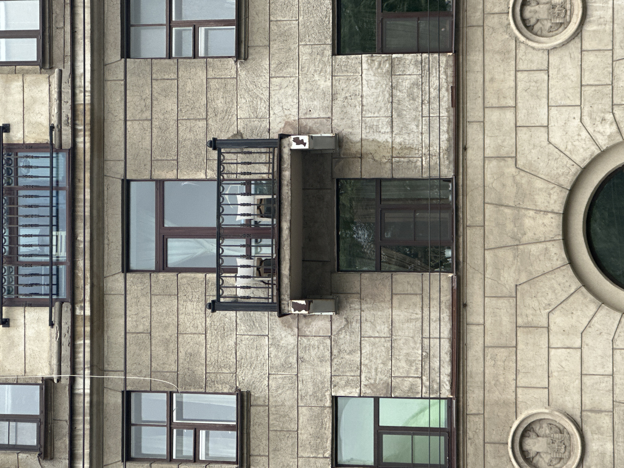 The image shows a facade of a historic apartment building in Saint Petersburg, Russia. The building is constructed of light-colored stone blocks. The facade features several windows, a small balcony, and decorative architectural elements.
The windows are of various sizes and configurations, most with dark brown frames and panes of glass that reflect the overcast sky. Some windows have sheer white curtains visible.
A narrow balcony with a black metal railing is centrally located on the middle level. Two chairs and a small table are visible on the balcony, along with a light-colored cushion on one of the chairs. The balcony appears to be supported by a stone structure that shows signs of wear and staining.
Below the balcony, on the lower level, are two more windows. The left window is large and tall, while the right window is of a more standard size.
The lower section of the facade reveals a large, arched opening, partially visible at the bottom of the frame, which likely leads to an entrance. Two circular bas-reliefs are mounted on the facade to the left and right of this archway. These appear to be ornate carvings, though their specific subject matter is indistinct due to the resolution and distance.
The overall impression is that of an older building, possibly from the late 19th or early 20th century, typical of the architectural style found in many historic districts of Saint Petersburg. The sky appears overcast, suggesting a cloudy day. There are no people visible in the image.