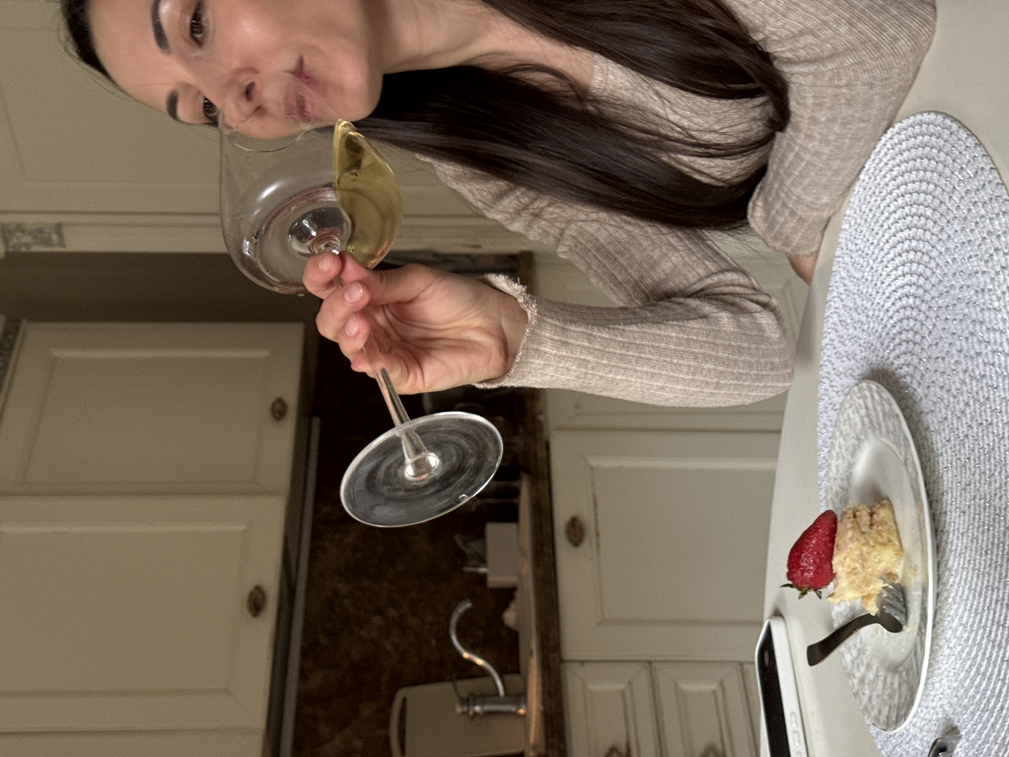 A woman with long, dark hair is holding a wine glass filled with a yellow liquid, possibly white wine, to her lips. She is wearing a light brown ribbed sweater. To her right is a plate with a piece of cake topped with a strawberry, and a fork. The plate is sitting on a circular gray placemat. In the background are white kitchen cabinets and a brown countertop with a faucet. The location in the city of Krasnoyarsk, Russia is not discernible from the image.