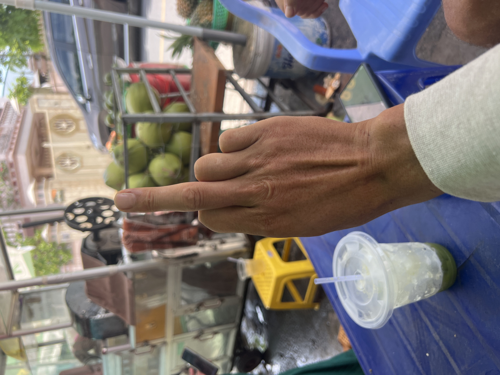 A person's hand, displaying a middle finger, is prominently featured in the foreground of the image. The hand appears to be resting on a blue plastic table. To the left of the hand is a clear plastic cup with a white lid and a straw, containing a green liquid with ice. In the background, there is a street scene with a cart laden with coconuts. The environment suggests an outdoor setting, possibly a market or roadside vendor. The specific location is identified as Binh Thuan Ward, Vietnam.