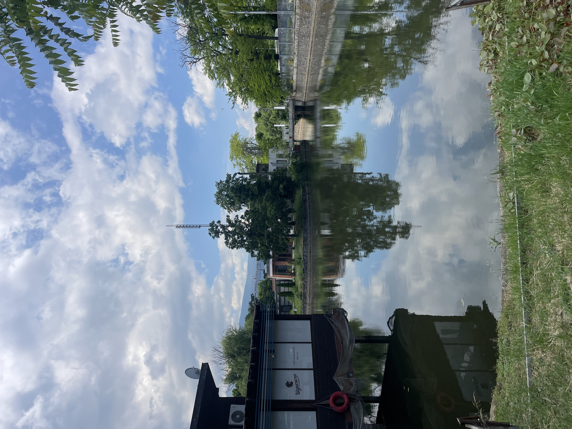 This image depicts a serene canal or river scene in Sofia, Bulgaria, on a clear, sunny day. The sky is a vibrant blue, filled with fluffy white cumulus clouds, partially framed by the green leaves of an overhead tree in the upper left corner.
On the left bank, a dark-colored building, possibly a restaurant or floating bar, extends into the water. Its facade features large windows, which display white text. Visible words include "Шуменско" (Shumensko), a Bulgarian beer brand, and "ЛЕГЕНДАРЕН ВКУС" (Legendary Taste), indicating a food or beverage establishment. An air conditioning unit is mounted on the side of the building, and a small satellite dish is on its roof. Decorative fishing nets hang from the structure, and a red lifebuoy is attached to its exterior, emphasizing its waterside location.
The calm water of the canal perfectly mirrors the sky and clouds above, creating a striking reflection that duplicates the atmospheric conditions. The reflections also show the inverted forms of trees and structures from both banks.
The opposite bank, on the right side of the frame, is covered in lush green grass and dense trees, with glimpses of distant buildings and what appears to be an elevated structure or bridge in the background. The overall impression is one of tranquility and natural beauty within an urban or semi-urban setting, with no people visible in the scene.