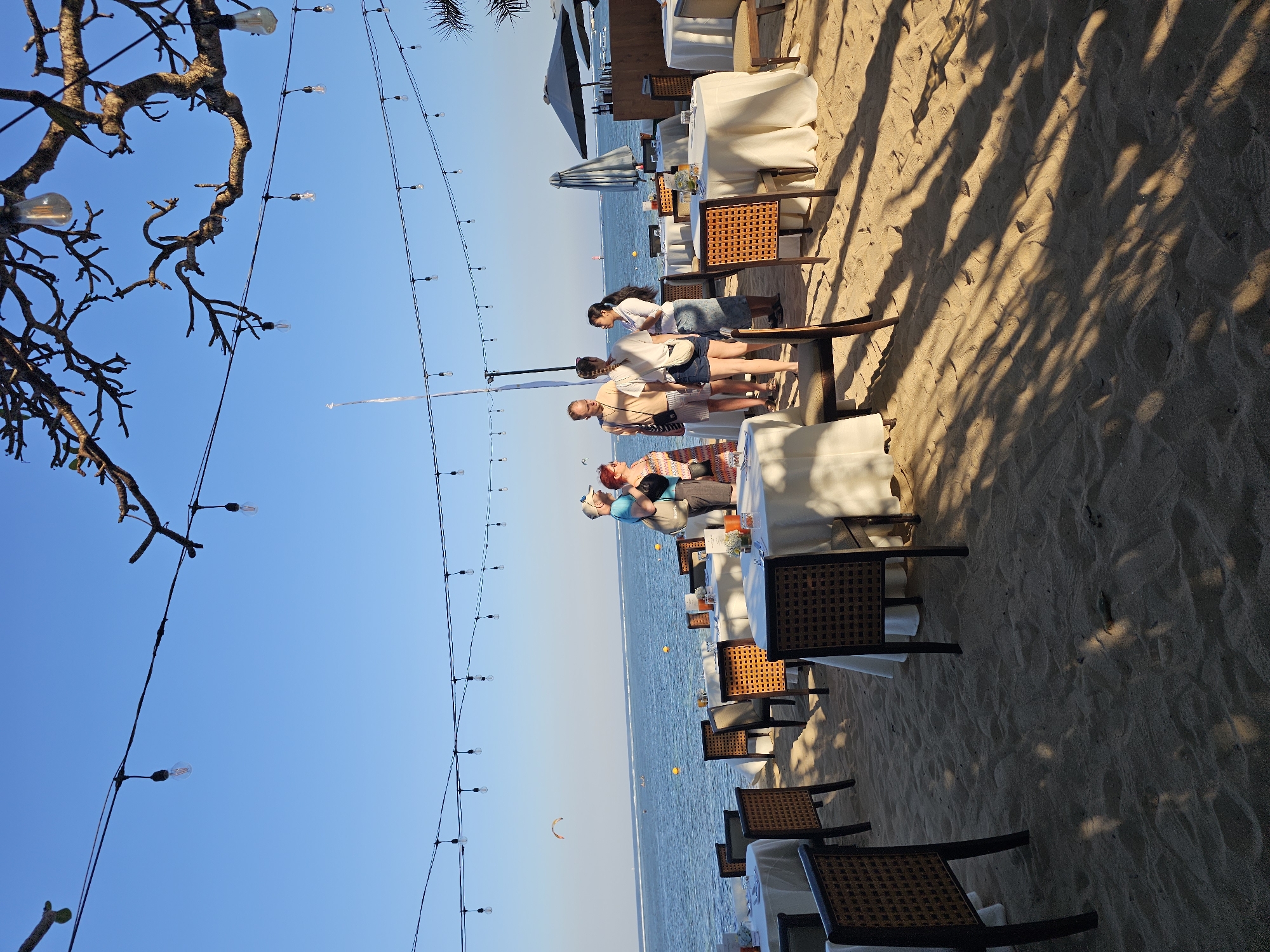 The image depicts a beachside dining area in Denpasar, Indonesia, likely a restaurant or event space on a beach. Set up on the sand are several round tables draped with white tablecloths, each surrounded by woven-back chairs. Overhead, strings of incandescent bulbs are strung across wires, casting a warm glow. In the background, the ocean is visible, with a clear blue sky above. A few people are gathered near some of the tables, appearing to be guests or staff. One man in a blue shirt and baseball cap, and a woman in a patterned dress are standing and conversing. Another group of four individuals, two men and two women, are also gathered in conversation near a table. In the distance on the water, a kiteboarder is visible, suggesting recreational activity. The lighting and setup indicate a pleasant, possibly evening, atmosphere.