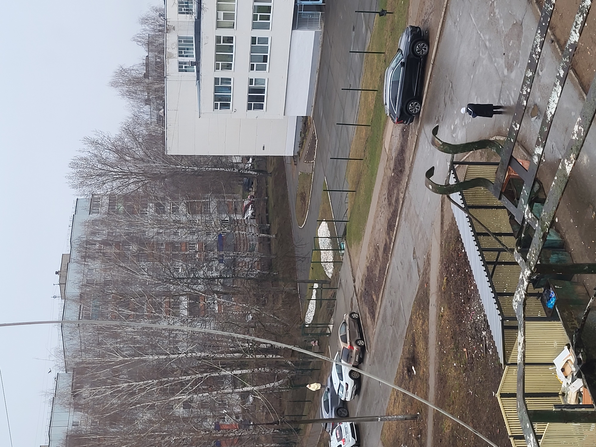 The image shows an aerial view of an urban scene, seemingly taken from a high vantage point in Chaikovsky, Russia. A tall white building with numerous windows dominates the upper left portion of the frame. Several bare trees indicate that it is likely a colder season. A street runs alongside the building, with several parked cars, mainly in shades of white and gray. A dark-colored SUV is parked closer to the top of the image. A person dressed in dark clothing with a light-colored head covering is walking on the sidewalk next to the SUV. In the right foreground is a weathered metal structure, possibly part of a balcony or building framework. Below the white building are additional apartment complexes visible in the background through the trees. The ground has patches of snow in places.