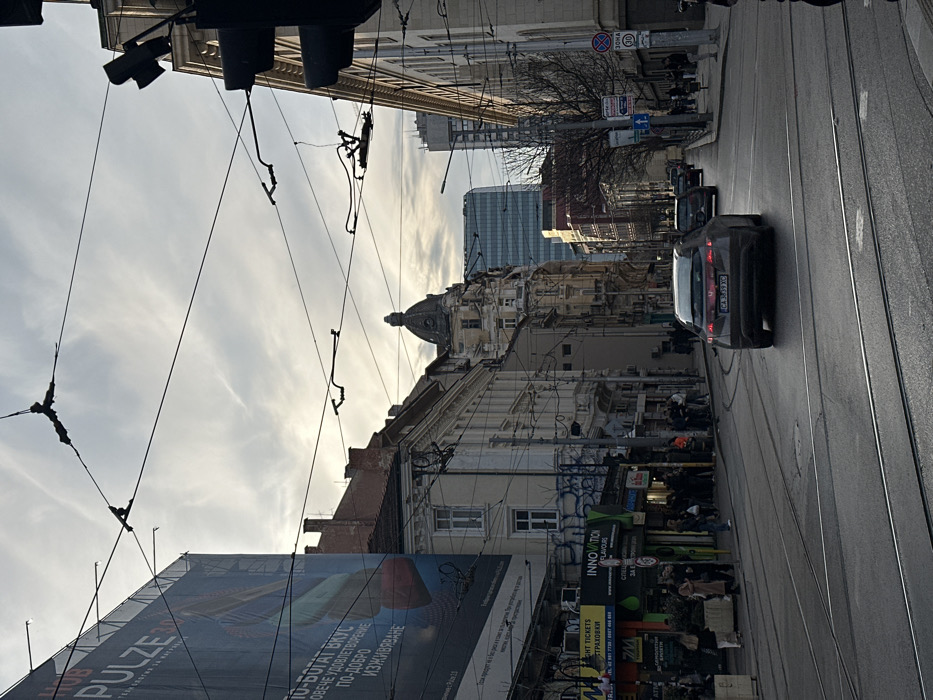 The image depicts Graf Ignatiev Street in Sofia, Bulgaria, viewed under an overcast sky. A dark grey hatchback, bearing Bulgarian license plate "CA 3839 XC", travels away from the viewer on a damp, asphalt road containing tram tracks. Other vehicles follow further down the street.
On the immediate left, a large billboard advertising "PULZE" with Bulgarian text ("ПО-БОГАТ ВКУС / ПОВЕЧЕ УДОВЛЕТВОРЕНИЕ / ПО-ДОБРО ИЗЖИВЯВАНЕ") covers a building facade. Below, storefronts such as "МОБИЛНИ ТЕЛЕФОНИ" and "INNOVATION FLAVOURS" are visible, with pedestrians congregating on the sidewalk. The buildings on this side are multi-story, older European architecture, some with visible graffiti.
On the right, a line of classical, ornate buildings extends into the distance. A prominent building with a distinctive domed roof and turreted features is visible at 10 Graf Ignatiev Street. Beyond these structures, modern glass skyscrapers are discernable in the background. A dense network of black overhead tram wires and support cables spans the sky. Pedestrians in winter attire populate both sidewalks. Roadside elements on the right include a 30 km/h speed limit sign and bare trees. Traffic lights are partially visible on the far right.