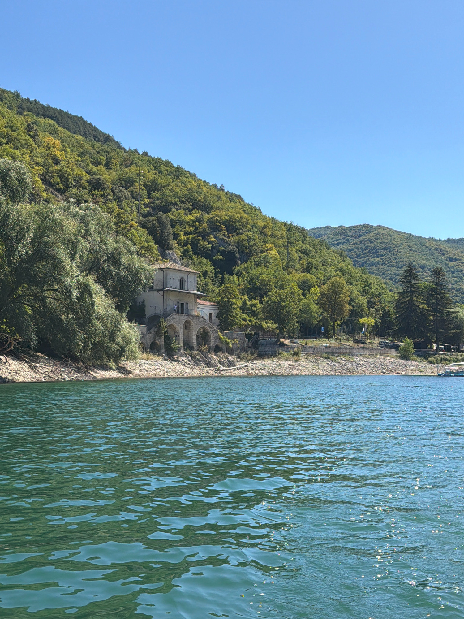 This is a daytime, eye-level, medium shot of a lake with a wooded hillside and a building in the background, taken from the city of Scanno, Italy. The lake occupies the bottom half of the frame, with the water appearing green and rippled under a clear blue sky. The shoreline is rocky and lined with trees. On the hillside, there is a building with arched windows and a balcony. The hillside is densely covered with green trees. The scene depicts a natural environment with a historical structure.