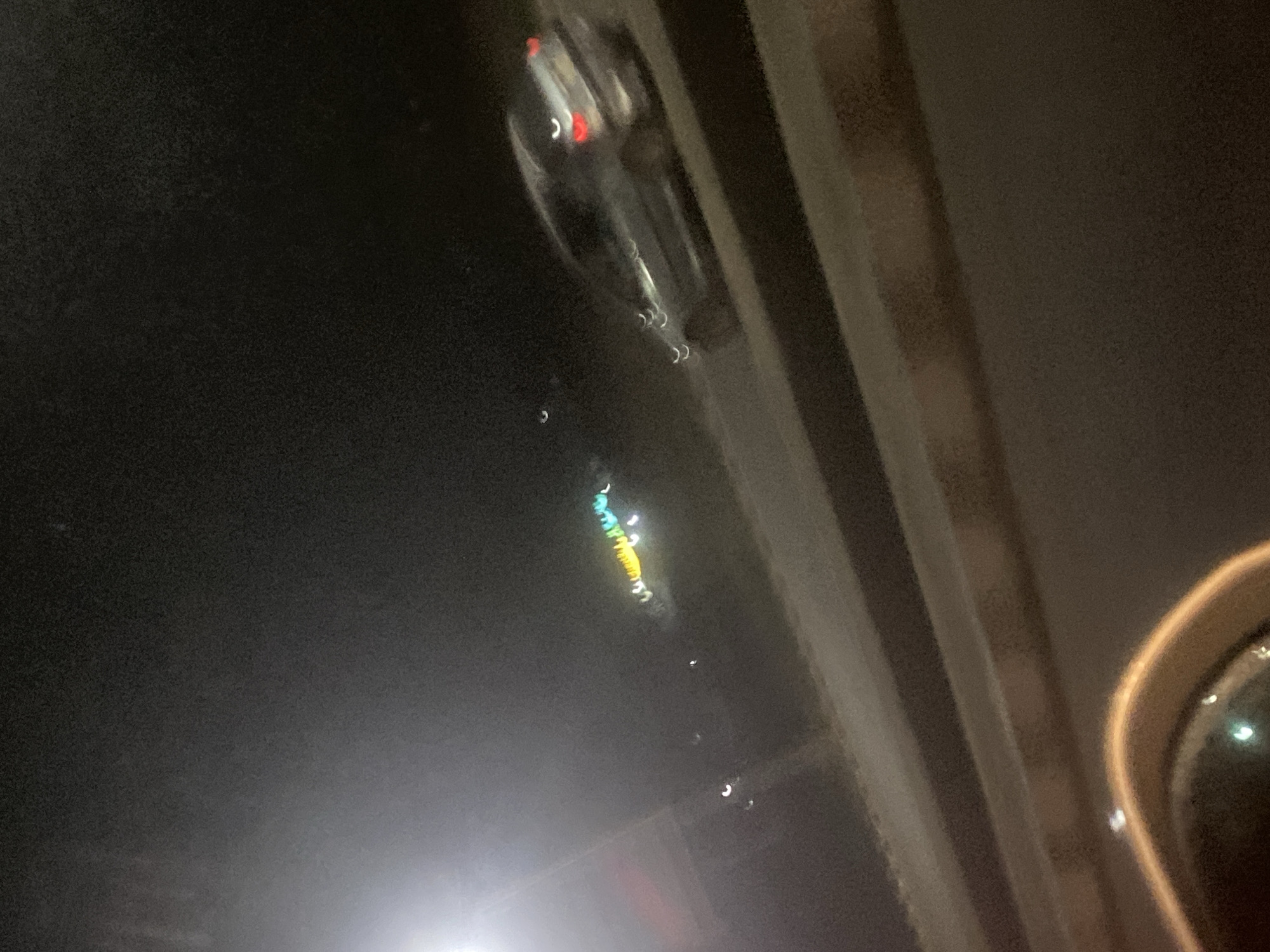 The image captures a night scene in Gosa, Nigeria, likely from a moving vehicle, as evidenced by the motion blur. In the mid-ground, a silver sedan is visible driving on a road. The vehicle's taillights are illuminated red, indicating its presence and direction of travel. Further in the distance, a blurry, elongated streak of multi-colored lights suggests a sign or structure, possibly a business or advertisement, with colors like yellow, green, and blue. The background is dark and indistinct, typical of a nighttime urban or semi-urban environment. The foreground shows the interior of a vehicle, with a portion of a window frame or dashboard visible. The overall impression is one of a transient moment, capturing a glimpse of the city at night.