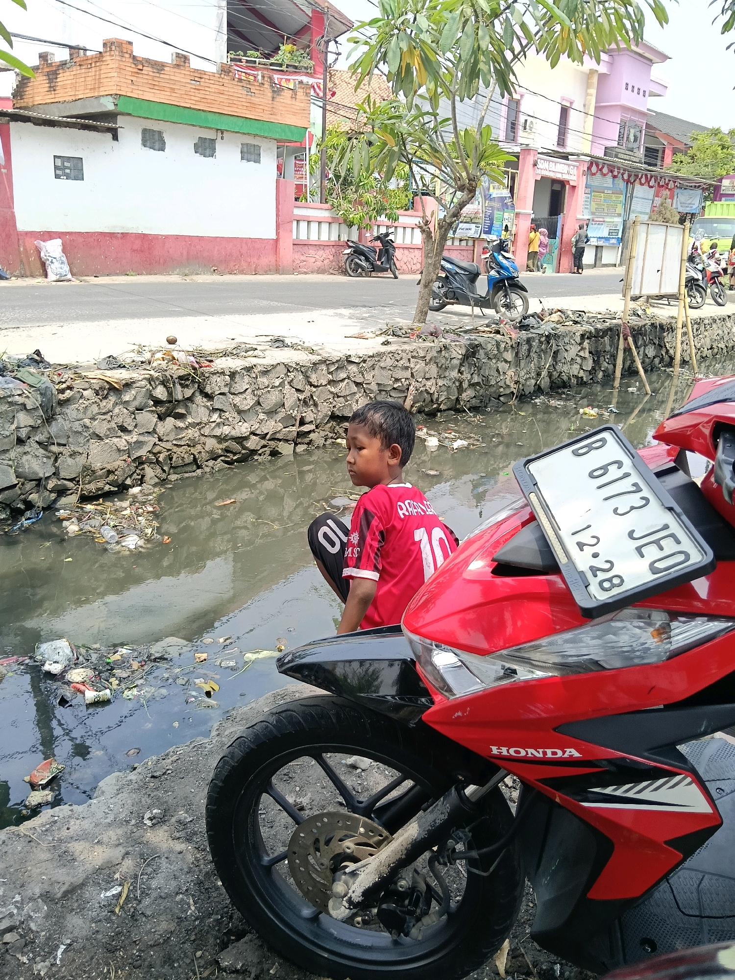 In the city of Belimbing, Indonesia, a young boy in a red and black jersey sits on a red motorcycle. The boy's jersey has the number 10 on the back. The motorcycle is parked on a dirt embankment next to a polluted canal filled with garbage. The canal has a stone wall lining its edge. In the background, buildings line a street, and other motorcycles are visible. The overall scene depicts a somewhat neglected urban environment.