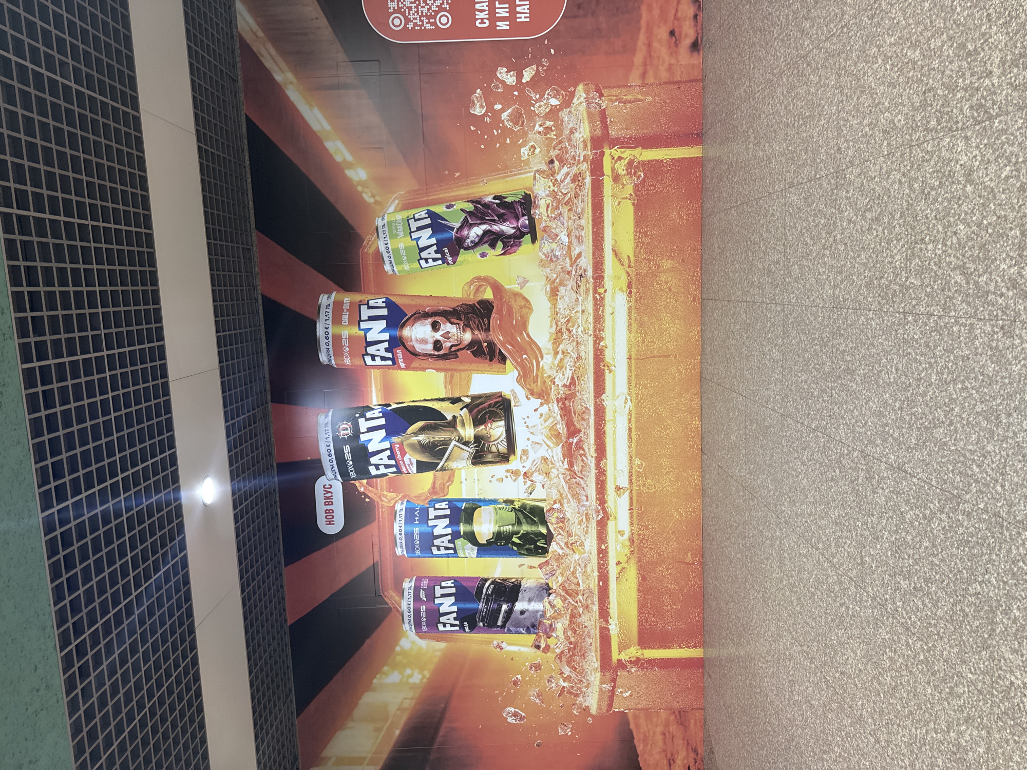 The image shows a promotional poster for Fanta soda, featuring various cans with video game character designs, affixed to a wall. The poster is advertising a promotion in partnership with Xbox, celebrating 25 years of the brand, as indicated by the "XBOX 25" text and logos next to the Fanta brand name on each can. The cans feature characters from popular video game franchises such as Call of Duty, Warcraft, and Halo. The poster is positioned on a wall with a checkered tile design on one side and a plain, textured wall on the other. The promotional poster is written in Bulgarian, showing that the location of the image is most likely within the city of Sofia, Bulgaria.