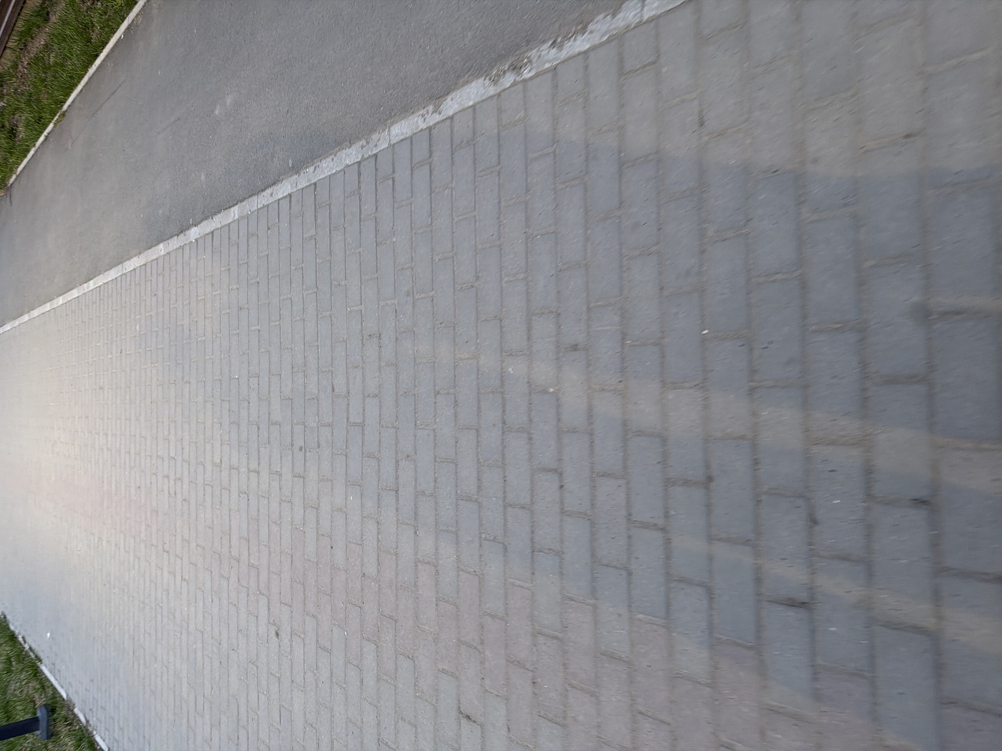 This image captures an outdoor scene featuring a paved pathway running parallel to an asphalt road. The pathway, which takes up the majority of the frame, is constructed from rectangular grey paving stones laid in a herringbone pattern. These stones exhibit variations in shade, suggesting texture and possibly slight wear.
A thin, somewhat uneven white line delineates the boundary between the paved path and the adjacent darker grey asphalt road. To the far left of the asphalt road, a strip of green grass is visible, suggesting a verge or embankment. A small, dark, unidentifiable object, possibly a post, is partially visible in the bottom left corner near the grass.
The image is captured in daylight, with some subtle shadows and lighting variations across the paved surface, indicating natural light conditions. No people or specific activities are visible. The overall impression is a quiet, functional pedestrian area alongside a road in what appears to be an urban or suburban setting. The image is oriented vertically, looking down the length of the path and road. There is no visible text in the image. The location is Chaikovsky, Russia.