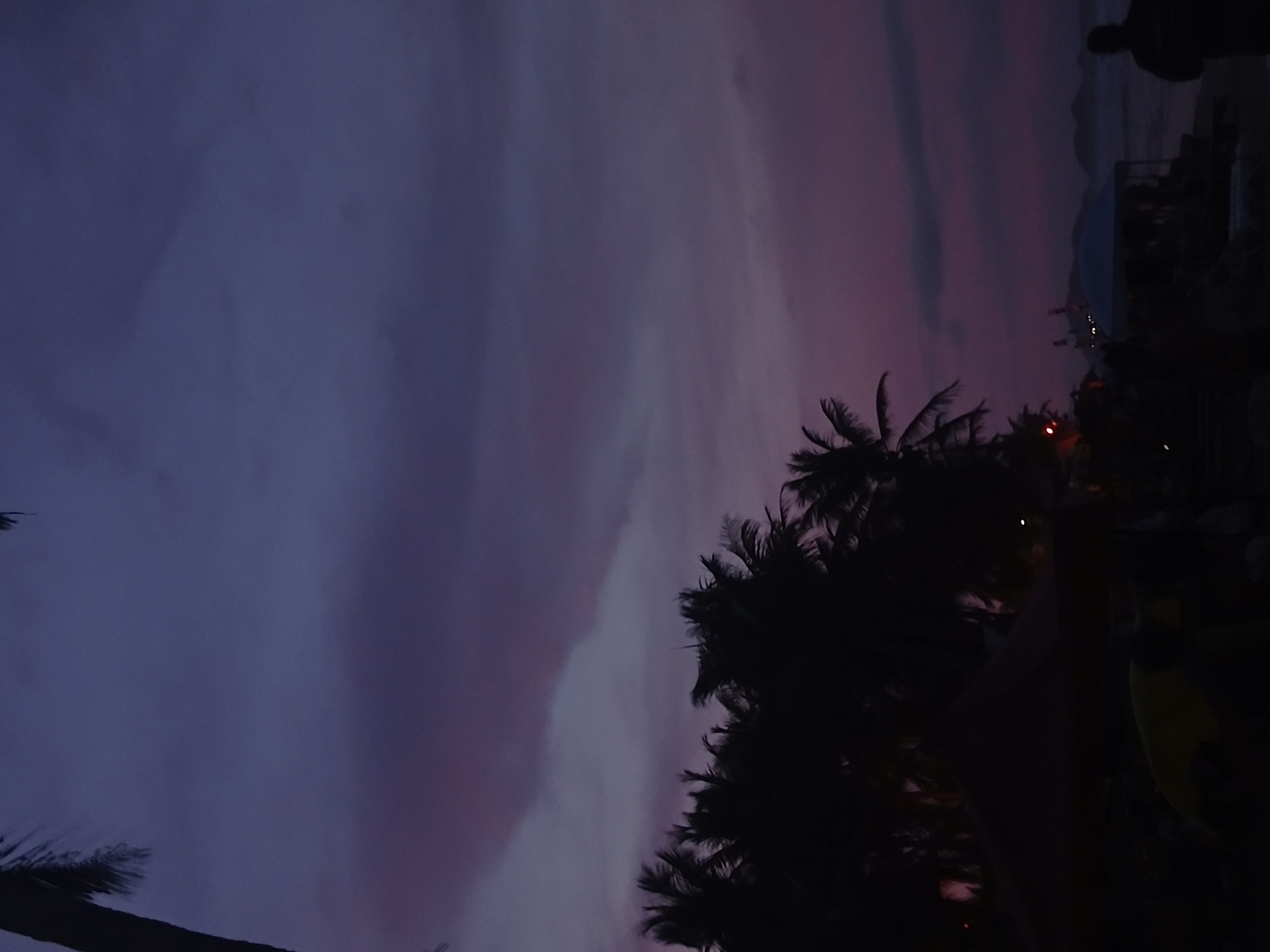 The image shows a dusk or dawn scene in Playa Parguito, Venezuela. The sky is a mix of dark blue and purple hues. Silhouetted palm trees are visible in the lower right, indicating a tropical or coastal location. There is a building with multiple stories and some people near the building. The image has a low light, which suggests the time of day is late evening or early morning.