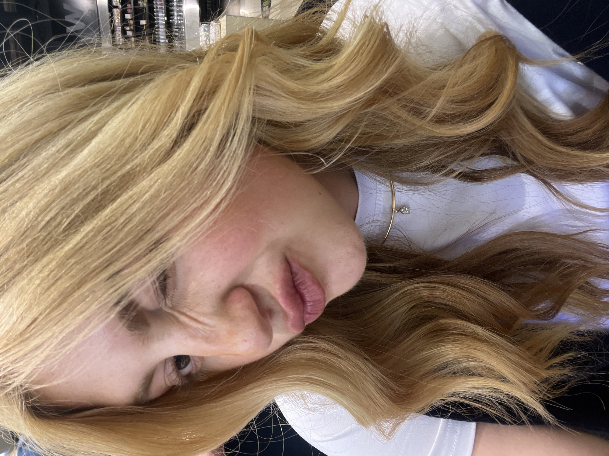 A close-up, slightly rotated image depicts a blonde woman with wavy hair. She is wearing a white t-shirt and a gold necklace with a small pendant. The background includes a dark area, possibly clothing, and a blurred shelf-like structure with metallic and reflective elements. The location in Johannesburg, South Africa, is not apparent from the visual information provided in the image.