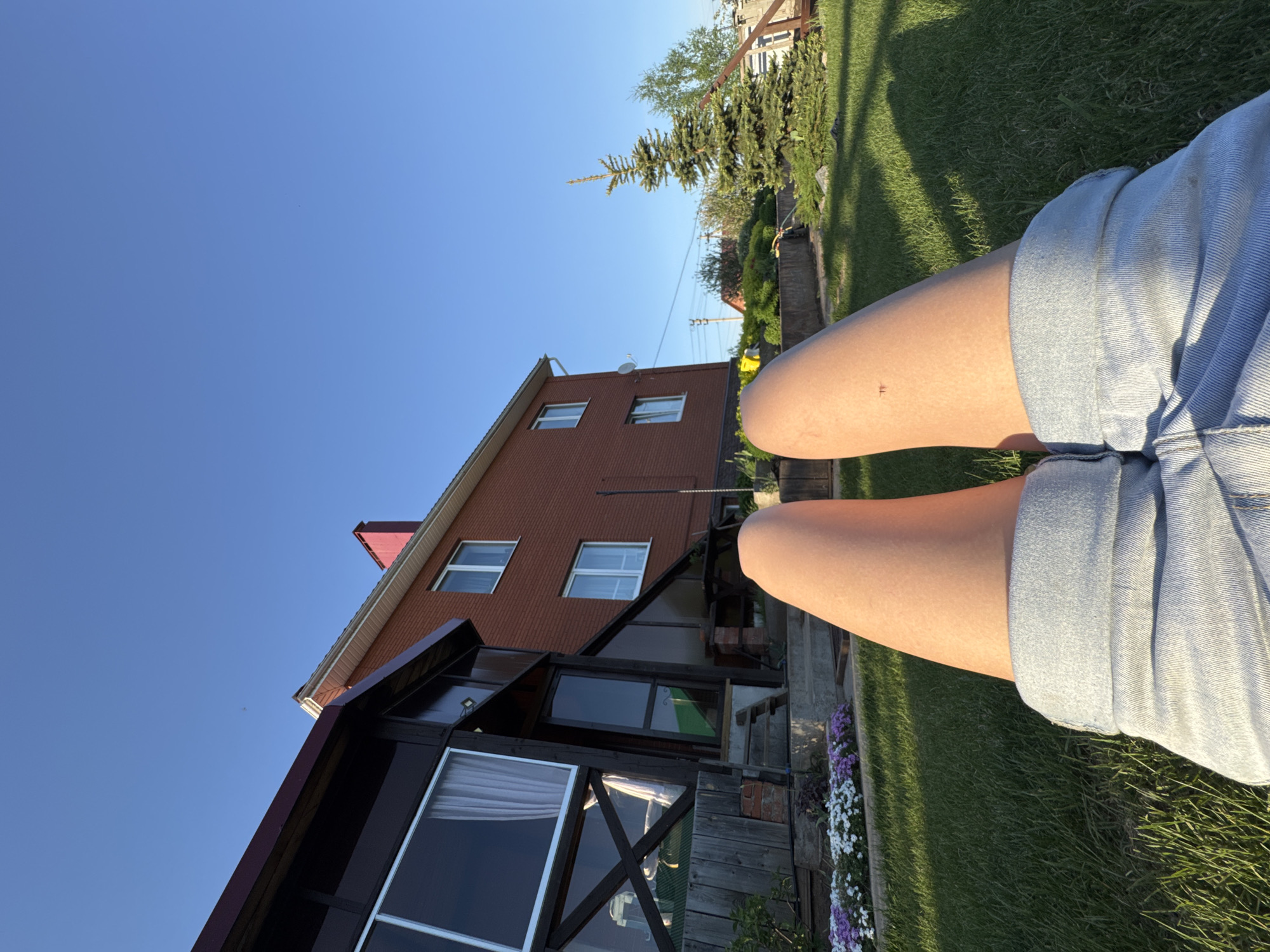 The image shows a view from a low angle, likely from a person sitting on the grass. In the foreground are the person's legs wearing denim shorts. The background reveals a two-story brick house with a dark-colored extension or porch featuring large windows. A patch of green lawn is visible, along with some flowers near the house. Above the house, the sky is clear blue. The location is Прикамский, Russia.