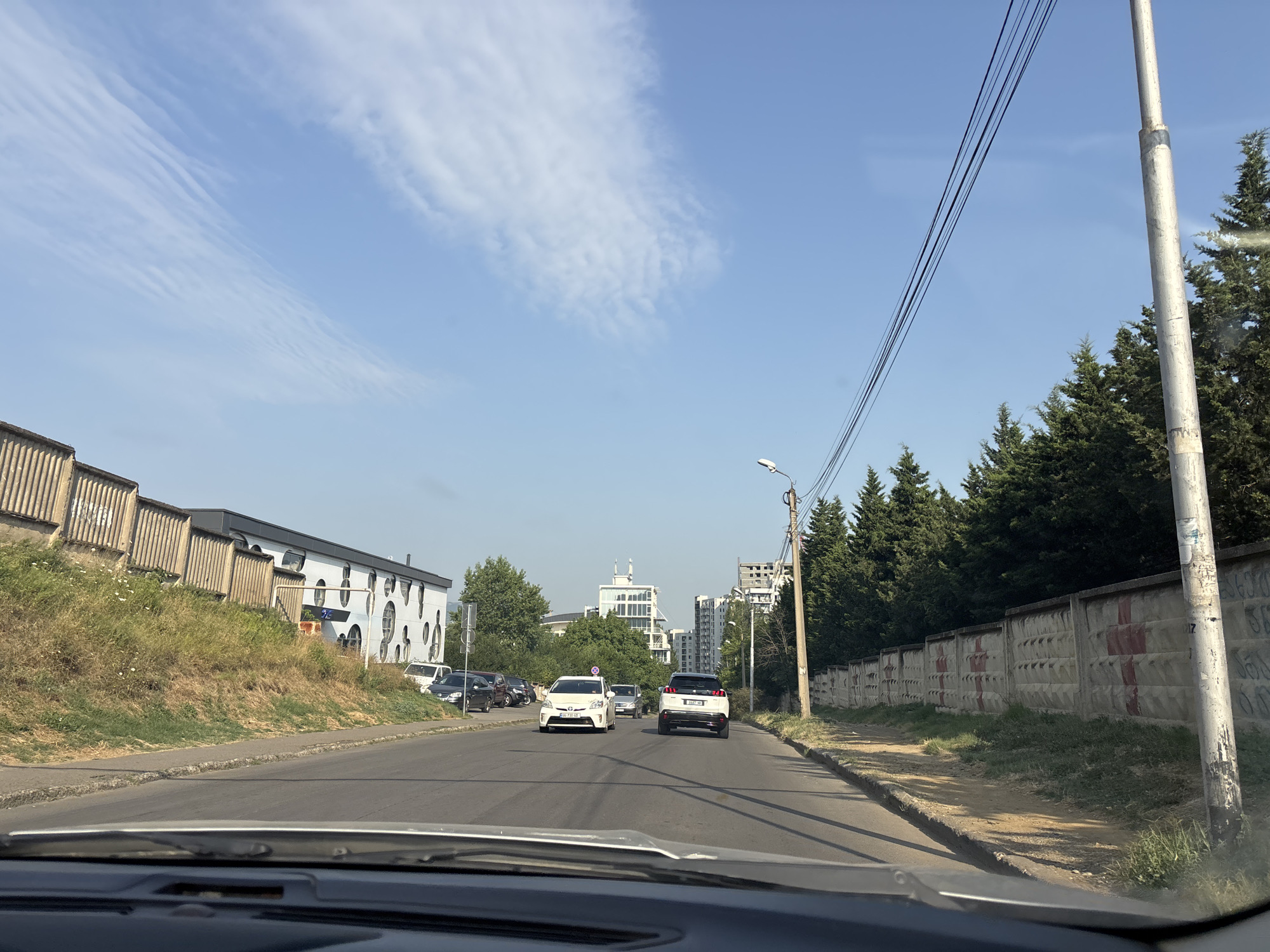 The image depicts a street scene in Tbilisi, Georgia, viewed from the perspective of a vehicle. The road is paved and appears to be a multi-lane street. On the left side of the road, there is a grassy embankment topped with a concrete structure with vertical supports. Further along the embankment, a white building with circular window-like openings is visible. On the right side of the road, a concrete wall with a repeating pattern of red crosses runs along the sidewalk, backed by a line of coniferous trees. A utility pole with power lines stretches across the upper right portion of the frame.
Several vehicles are present on the road. In the foreground, the hood and windshield wipers of the vehicle from which the photo is taken are visible. Ahead, a white car, possibly a Toyota Prius, is in the left lane. In the right lane, a white SUV, possibly a Peugeot 3008, is ahead of the Prius. Other cars are parked or moving further down the street. In the distance, several multi-story buildings, including a modern glass-fronted one, are visible, suggesting an urban or suburban environment. The sky is a clear blue with scattered wispy white clouds. The overall lighting suggests it is daytime, likely late morning or afternoon.