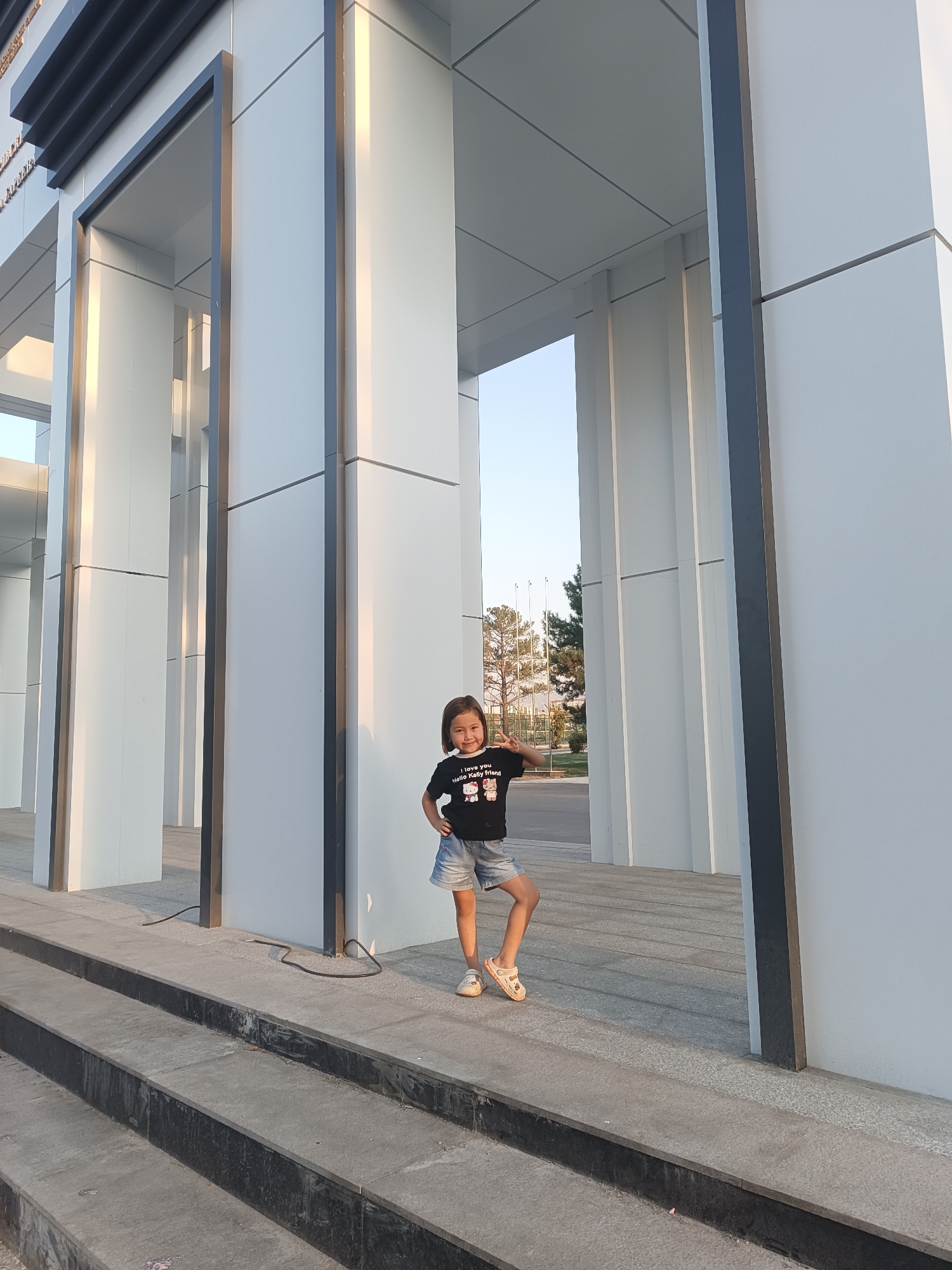 A young girl stands on a paved area in front of a modern, minimalist building. The building features large, white, rectangular pillars and dark gray framing around the openings. The girl is wearing a black t-shirt with cartoon characters, denim shorts, and light-colored sandals. She has her hands on her hips and is looking at the camera, with her other hand raised in a peace sign. The ground in front of the building is paved with large, light-colored tiles, and there are dark gray steps leading up to this area. In the background, trees and flagpoles are visible under a clear sky. The lighting suggests it is daytime, possibly late afternoon, with sunlight casting shadows and highlighting the architectural features. The scene appears to be an outdoor public space.