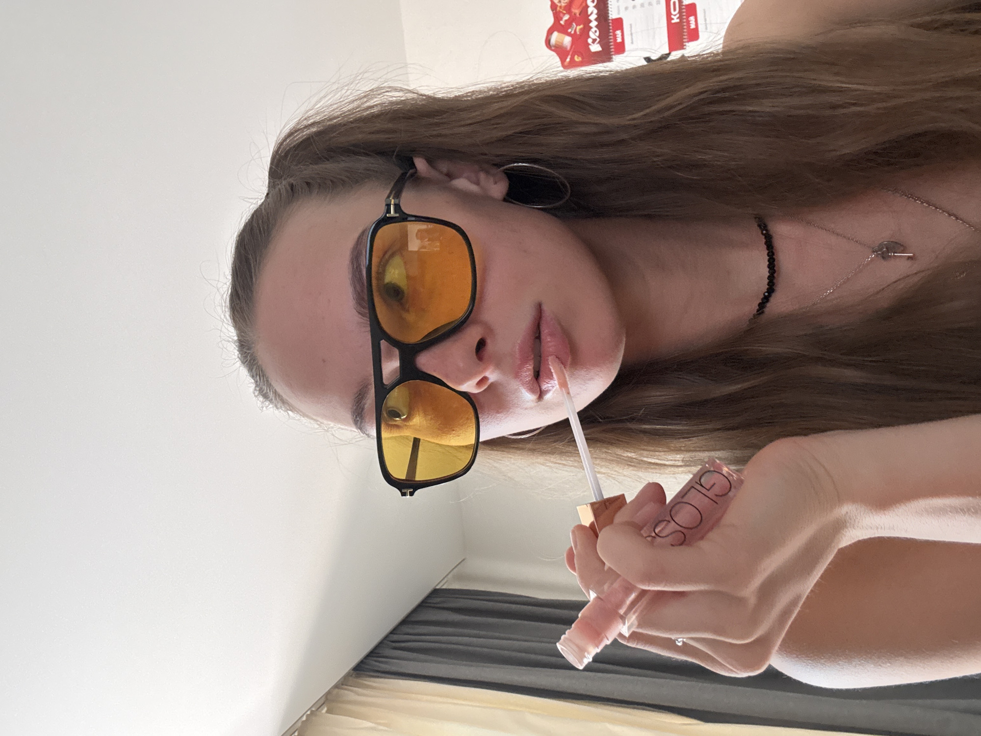 A young Caucasian female is applying lip gloss indoors, possibly in a bedroom. She has her hair in two pigtails and is wearing large, black-framed sunglasses with yellow lenses, hoop earrings, and a black beaded necklace with a silver key pendant. She is holding a clear tube of lip gloss with a pink hue, and the applicator wand is touching her lips. In the background, a gray curtain is visible to the left, and a calendar is hanging on the wall to the right, with Cyrillic text visible. The lighting appears to be natural, coming from an unseen window.