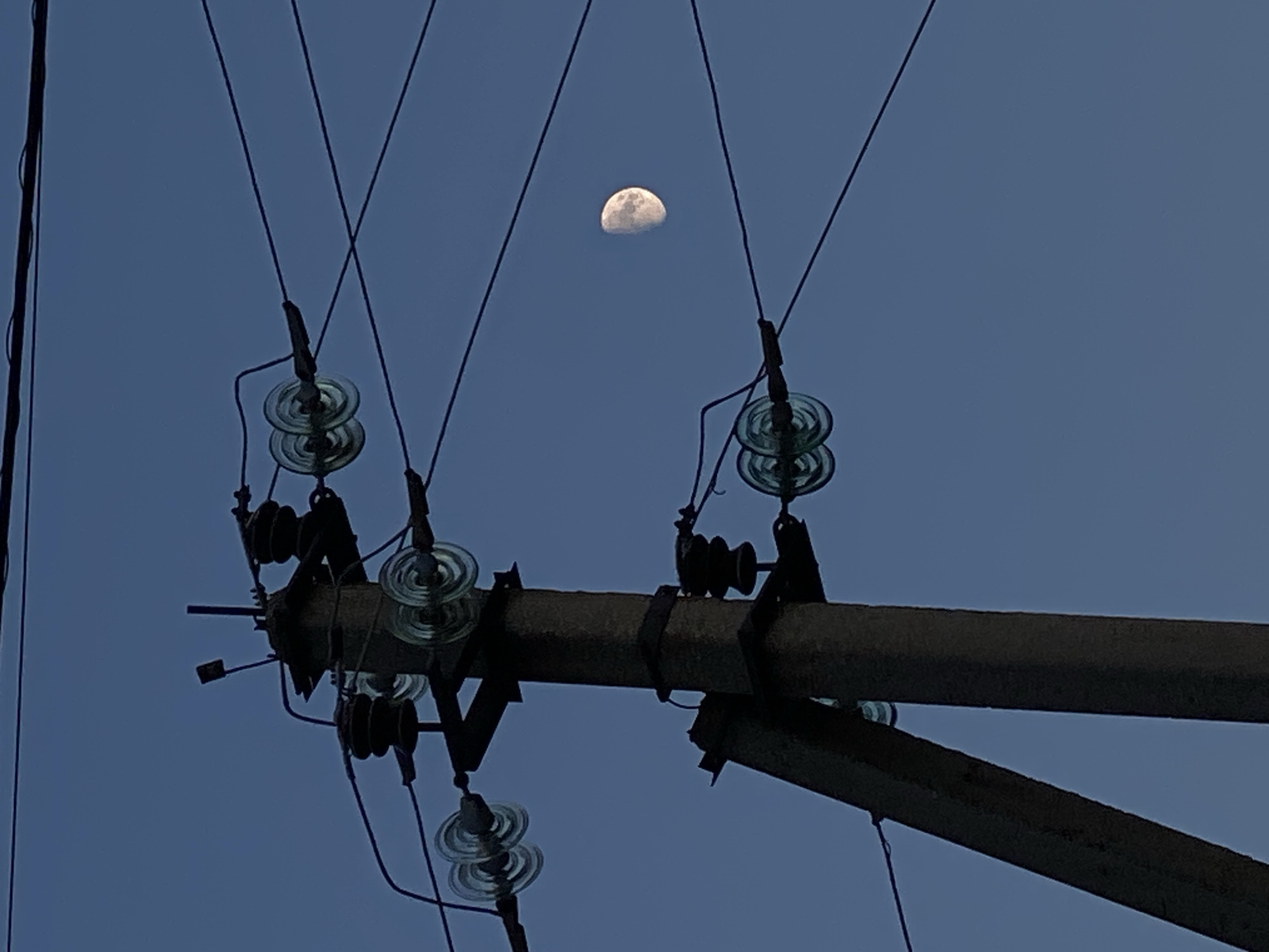 This is a low-angle, medium shot of a concrete utility pole supporting power lines against a clear, twilight blue sky. The moon, approximately half-illuminated, is visible in the upper right quadrant of the image. The utility pole is dark and appears to be weathered, with several insulators made of clear glass or plastic attached to it. These insulators hold taut black electrical wires that stretch across the frame. The overall scene is dimly lit, suggesting it was captured at dusk or dawn. The location is Cairaclia, Moldova.