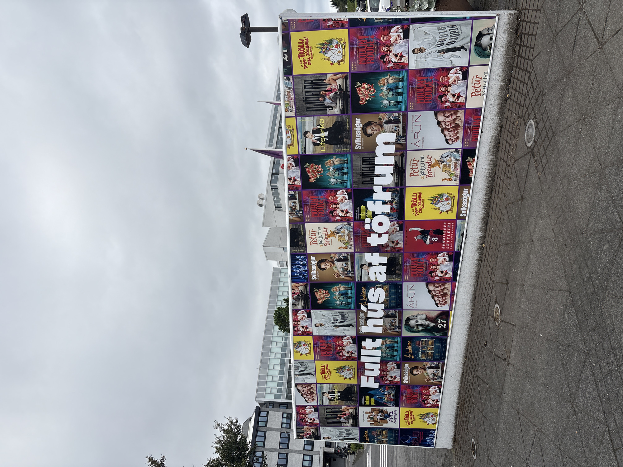 This is a medium-shot, eye-level outdoor photograph of a large billboard displaying numerous movie posters. The billboard is situated in front of a modern, grey building with a multi-paned glass facade. The sky is overcast and grey. The billboard features a central message in large white letters that reads "Fullt hús af töfrum." The individual movie posters are varied in color and content, showcasing actors and stylized graphics. The ground in front of the billboard is paved with grey interlocking stones and features several circular metal grates. To the left of the billboard, a portion of a dark green tree is visible. The scene appears to be in a public urban area, likely Reykjavik, Iceland, given the language on the billboard.