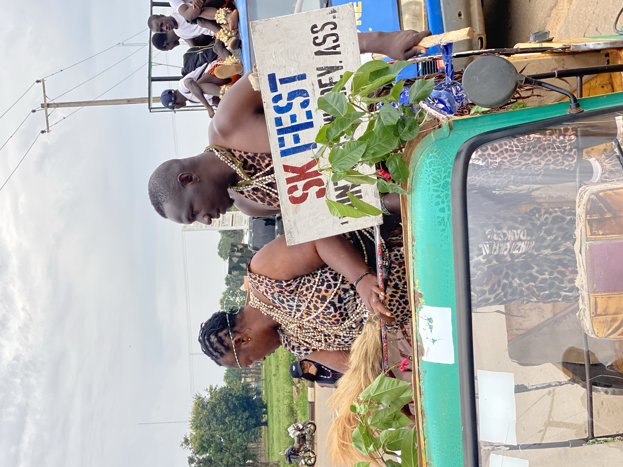 The image shows two adults wearing traditional clothing, adorned with necklaces of beads and cowrie shells, riding on what appears to be the back of an auto rickshaw. The man is holding a sign that reads "SK FEST" along with "AN DEV ASS." Several young people are sitting on a vehicle ahead of them. The vehicle also features decorative green plants and a leopard print covering the interior. The background consists of buildings, trees, and a partially overcast sky. A motorcycle is also seen in the background. The location is likely Maigizo, Nigeria, indicated by the sign.