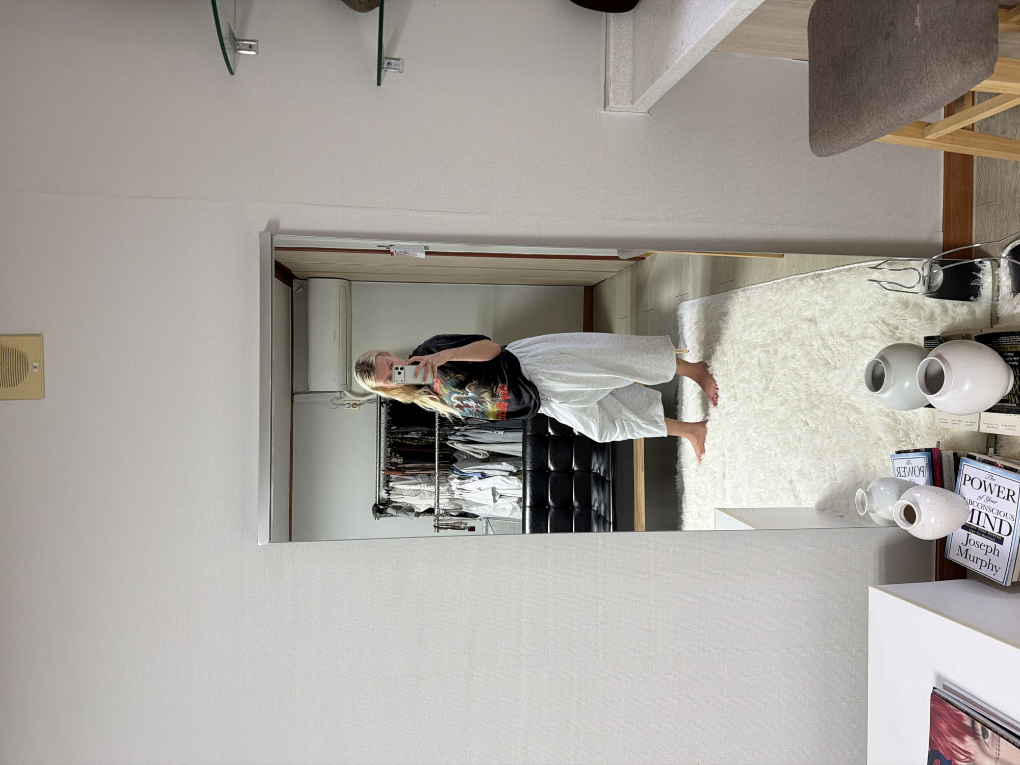 A woman is standing in front of a mirror, holding a phone and taking a picture of herself. The mirror reflects a closet with clothes hanging inside. There is a white rug on the floor near the mirror. On a white surface next to the rug are several vases and books, including "The Power of Your Subconscious Mind" by Joseph Murphy. The setting appears to be an interior space, possibly a room in an apartment or house. It is not possible to infer the location in Seoul, South Korea from the image.