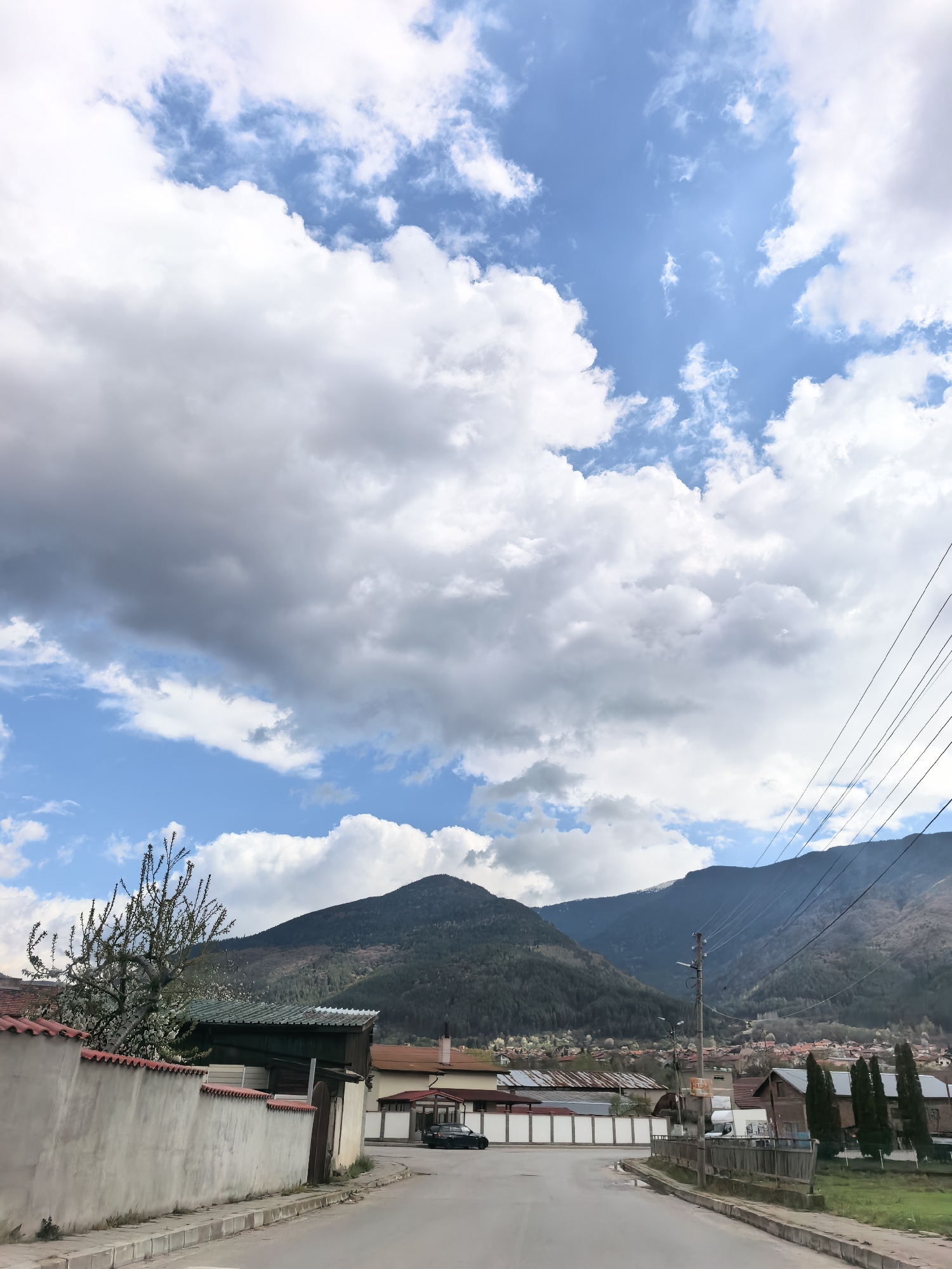 The image shows a street in an unknown city, with a view of mountains in the background. The sky is partly cloudy, with a mix of white and gray clouds. The street is empty and paved. On either side of the street, there are buildings, fences, trees, and power lines. A car is parked along the street. The overall scene is typical of a small town or residential area near a mountainous region.