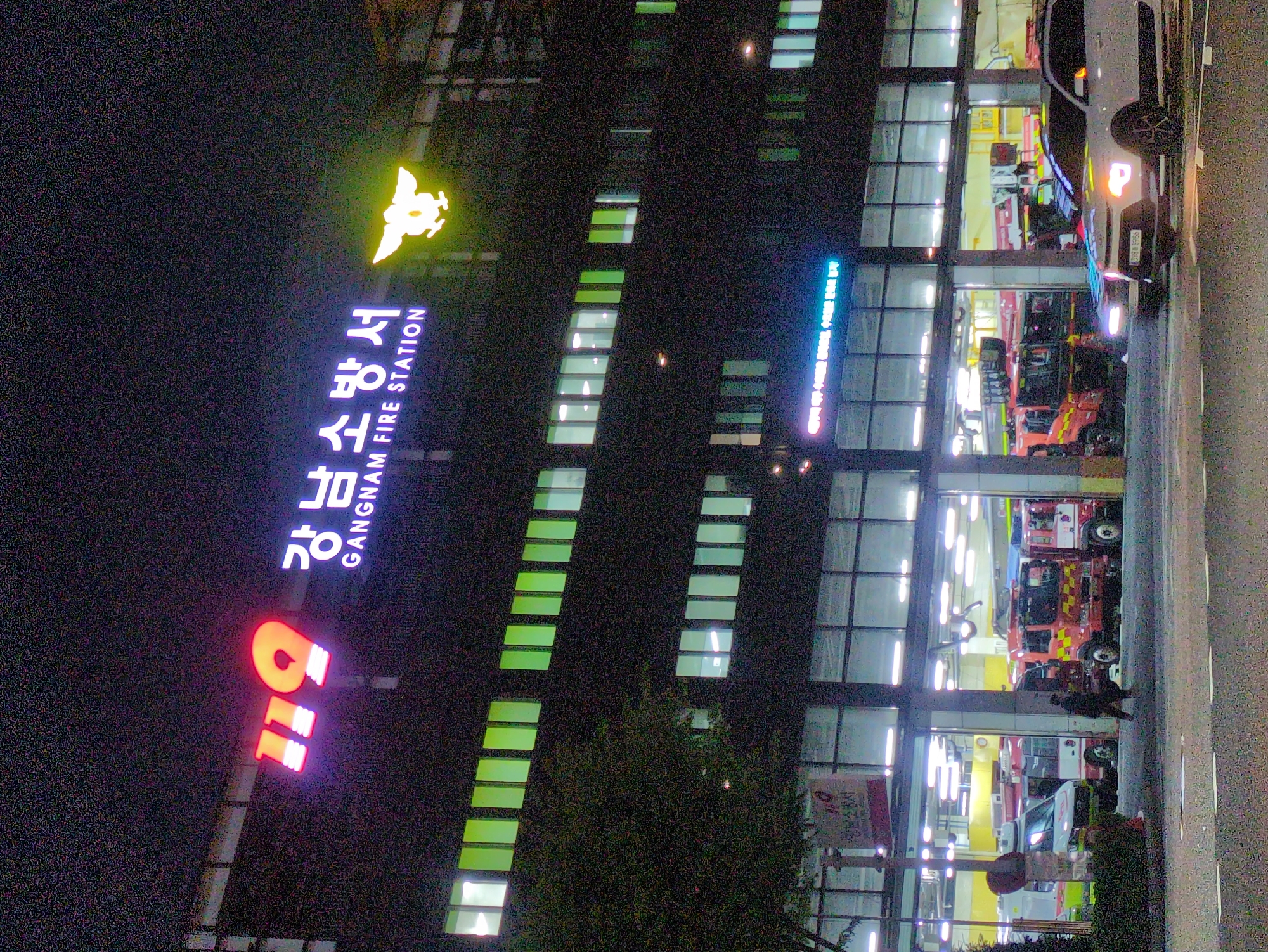 The image shows the Gangnam Fire Station in Seoul, South Korea, at night. The fire station's building is visible with its sign prominently displayed, along with the Korean number "119" in neon lights, indicating the emergency number. Rows of windows are illuminated. Fire engines and emergency vehicles can be seen inside the station's bays. A car is parked outside. People are present near the fire station's entrance. The environment is urban, illuminated by street and building lights.