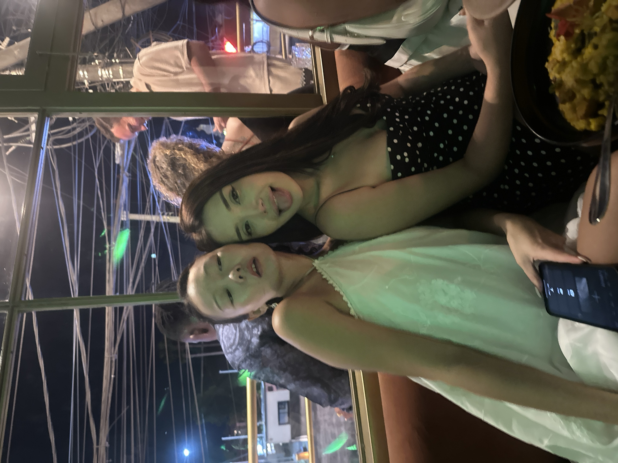 Two young women are posing for a photo inside a building or enclosed space. The woman in the foreground is wearing a white dress, and the woman behind her is wearing a black and white polka dot dress. Both are looking at the camera. A partially visible hand holds a cell phone. A plate of food is visible in the upper right corner. Outside the windows are building lights and architectural details that suggest the city of North Kuta, Indonesia. A man in a grey shirt can be seen in the background.