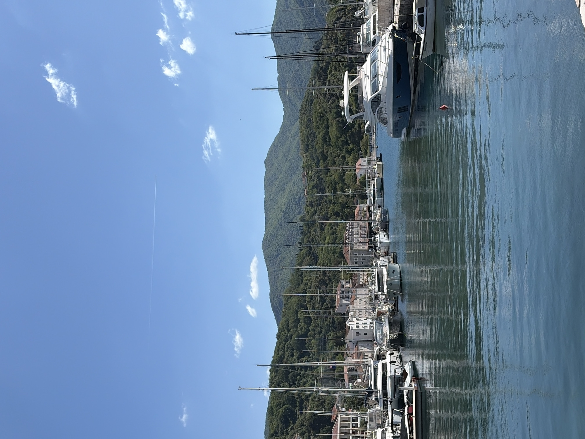 The image depicts a marina in Herceg Novi, Montenegro, filled with numerous boats and yachts docked along a waterfront. The water is calm, reflecting the boats and the adjacent coastal structures. In the background, a steep, verdant mountain rises, covered densely with trees. Buildings are nestled between the mountainside and the harbor, suggesting a settlement along the coast. The sky is clear blue, dotted with sparse clouds and a contrail from an airplane.