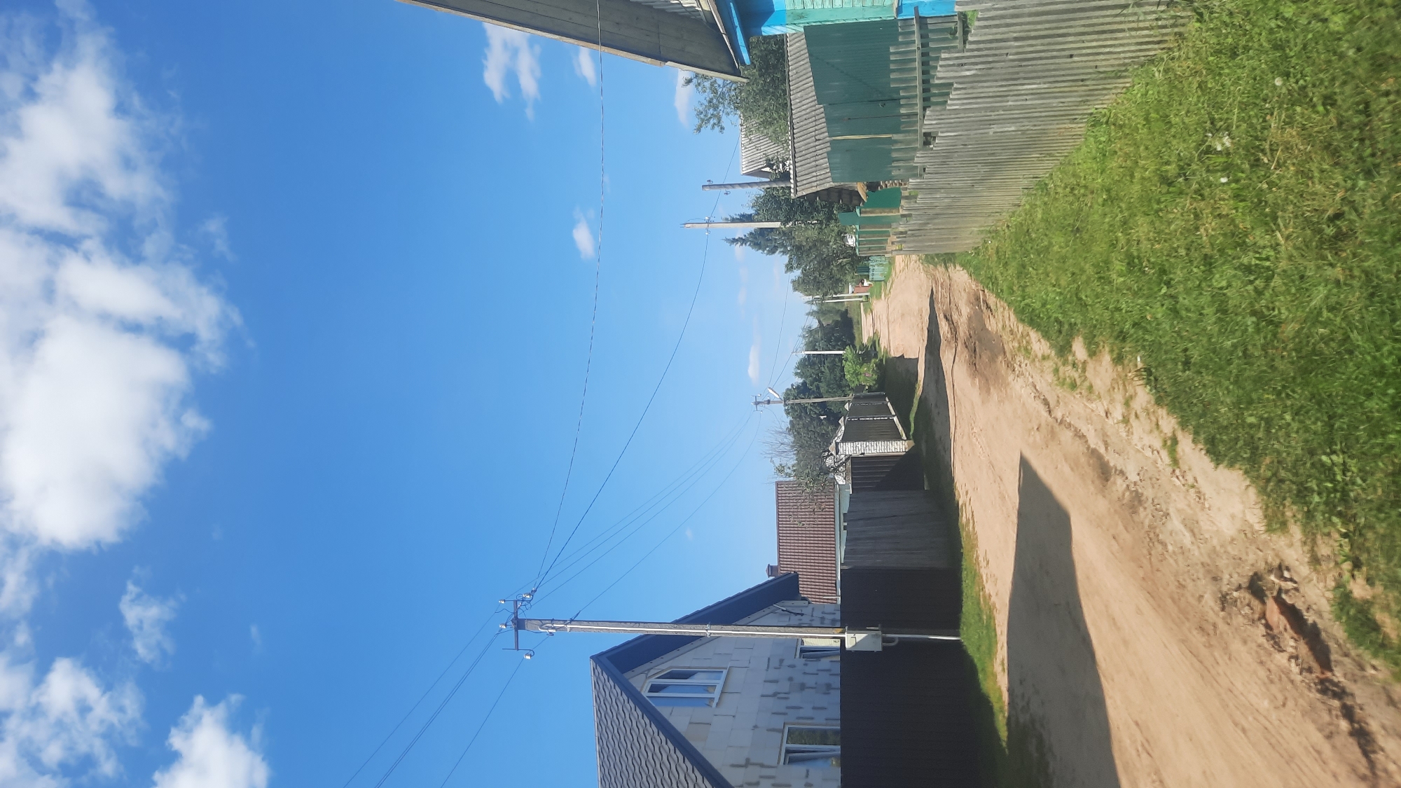 The image depicts a dirt road in a rural or suburban area, likely in or near the city of Shklov, Belarus. The road is unpaved and appears somewhat uneven with some minor damage. On the left side of the road, there is a light-colored house under construction, with exposed blockwork and a dark-colored roof. A dark fence runs along the property line. Further down the road on the left, there are more houses and fences, some with brick pillars. On the right side of the road, a light wooden fence encloses a grassy area. Beyond the fence, there are more houses and trees. Overhead, power lines are visible, supported by wooden utility poles. The sky is a clear bright blue with a few scattered white clouds, indicating fair weather. The overall impression is of a quiet, everyday scene in a residential area, possibly a village or a dacha community.