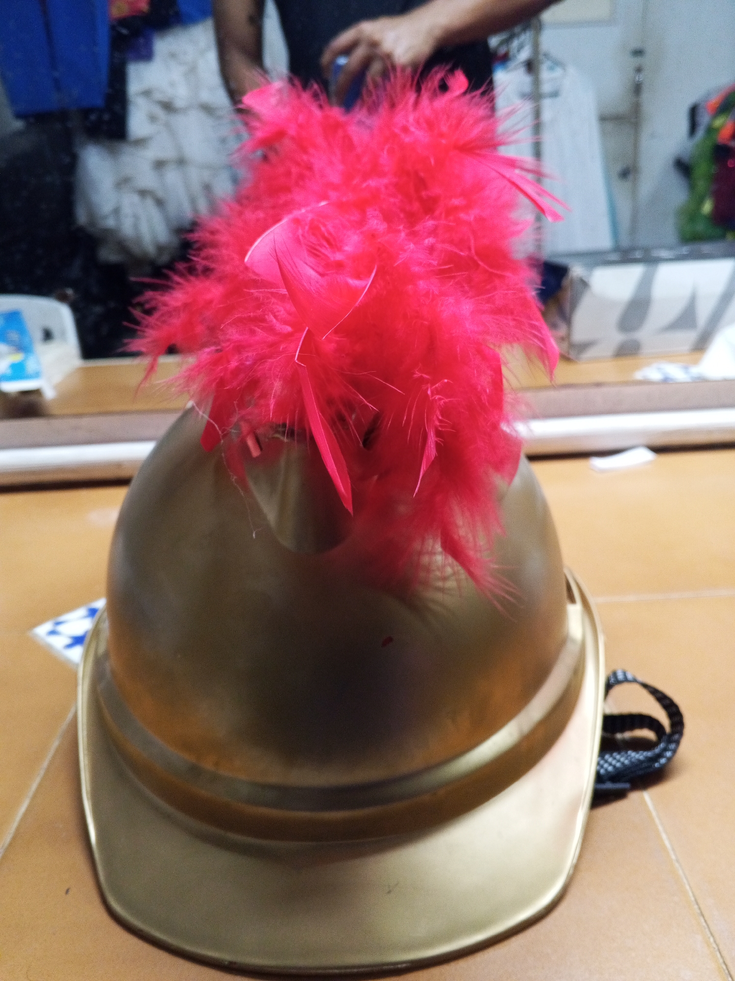 The central subject of the image is a golden-colored helmet, possibly a ceremonial or decorative item, adorned with a large, vibrant pink feather plume on top. The helmet has a metallic sheen, reflecting some of the ambient light. A black strap, likely a chin strap, is visible curled on the tiled floor to the right of the helmet.
The helmet is resting on a light brown or tan tiled floor. To the left of the helmet, a small piece of paper or a sticker with a blue and white patterned design is partially visible on the floor.
The background is out of focus but provides context for the scene. It appears to be an indoor setting, possibly a dressing room, storage area, or a room with a large mirror. Reflections of clothing, including a white, ruffled garment, are visible in what seems to be a mirror. A person, whose arm and torso are partially visible, can be seen in the blurred background, further suggesting an active space. The lighting indicates an indoor environment.
The image does not depict any specific activity but hints at preparation or storage related to costumes or performance attire in Porlamar, Venezuela. No discernible text is visible in the image.
