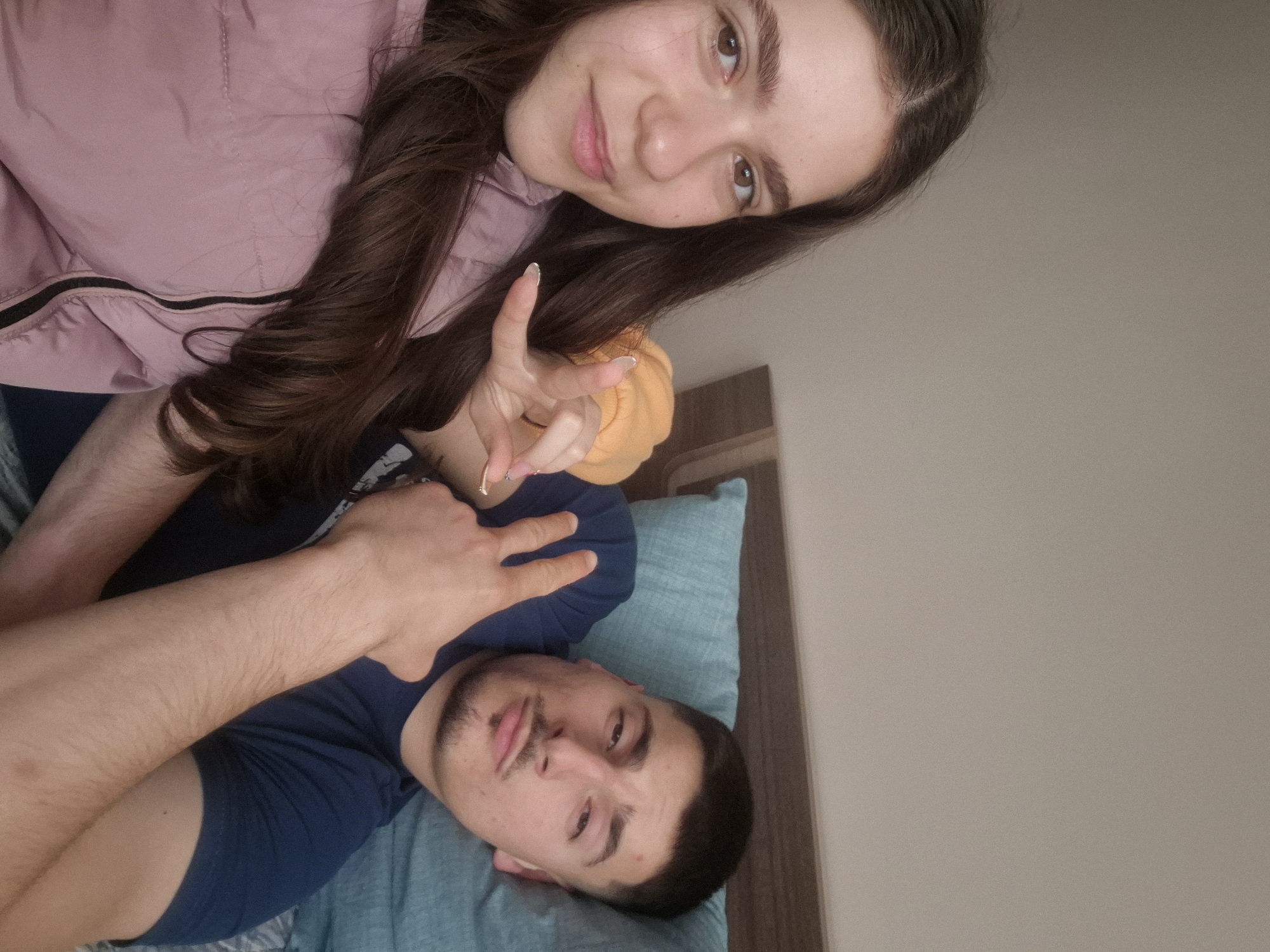 This media file presents a casual indoor selfie featuring two young individuals, a male and a female, situated in Pavlikeni, Bulgaria. Both subjects appear to be lying down, facing the camera with relaxed expressions.
In the upper portion of the image, a female with long, wavy dark brown hair and brown eyes displays a gentle smile. She is dressed in a light pink jacket and holds her left hand up, making a "V" or peace sign with white-tipped fingernails. Her long hair cascades around her shoulder and the jacket.
Below her, the male subject is visible, also looking towards the camera. He has short dark hair, a mustache, and some stubble on his chin. He wears a dark blue t-shirt and rests his head on a light blue pillow. His right hand is also raised, forming a "V" or peace sign.
The setting appears to be indoors, with a plain, light beige-colored wall forming the background, suggesting a bedroom or living area. A dark wooden furniture piece, possibly part of a bed frame or headboard, is partially visible on the right side. The overall impression is one of a relaxed, friendly moment shared between the two individuals. There is no visible text in the image.