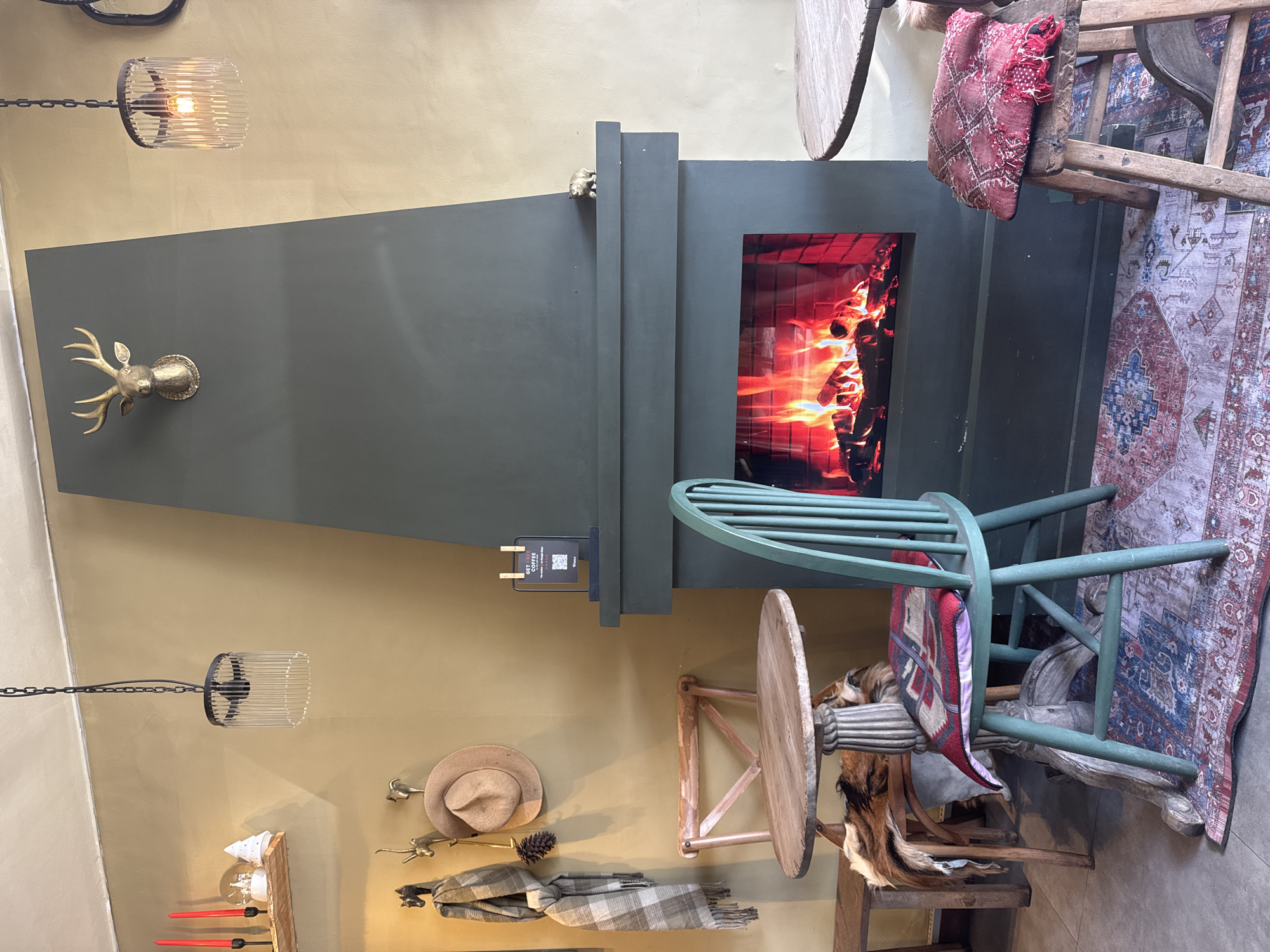 The image showcases the interior of a space, possibly a cafe or a shop, located in Unknown, Unknown. A tall, rectangular fireplace painted in a muted green tone is the focal point. Flames can be seen within the firebox. A golden deer head is mounted on the wall to the left of the fireplace. Above and below the fireplace, there are industrial-style light fixtures with cage-like shades. To the right of the fireplace, there is a collection of vintage wooden chairs, some draped with patterned textiles and pillows. A patterned rug is placed on the floor in the lower right corner of the frame. On the wall to the left of the fireplace, a hat and scarf are hanging, along with other decorative items on a wooden shelf.