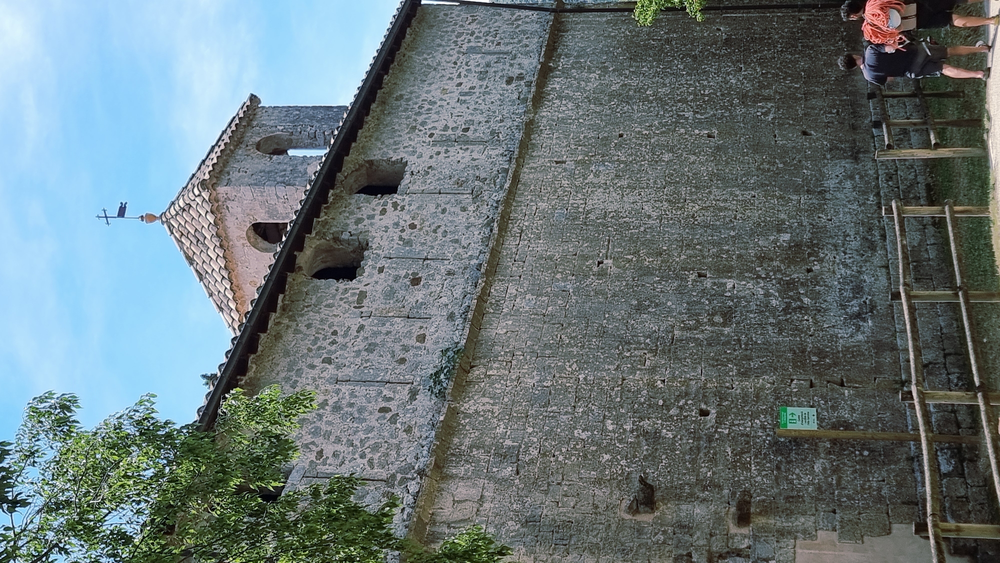 The image shows a stone building with a tiled roof and a bell tower topped with a cross. The building has a rustic appearance, with rough-hewn stone blocks and small, rectangular windows. In the foreground, a wooden fence runs along a path, and two people are visible walking away from the camera. One person is carrying a large coil of red rope over their shoulder. A green sign with white text and an arrow pointing right is mounted on a wooden post to the left of the fence. The sky is blue and clear, with some green foliage visible on the left side of the image. The scene appears to be outdoors, possibly in a rural or historical setting.