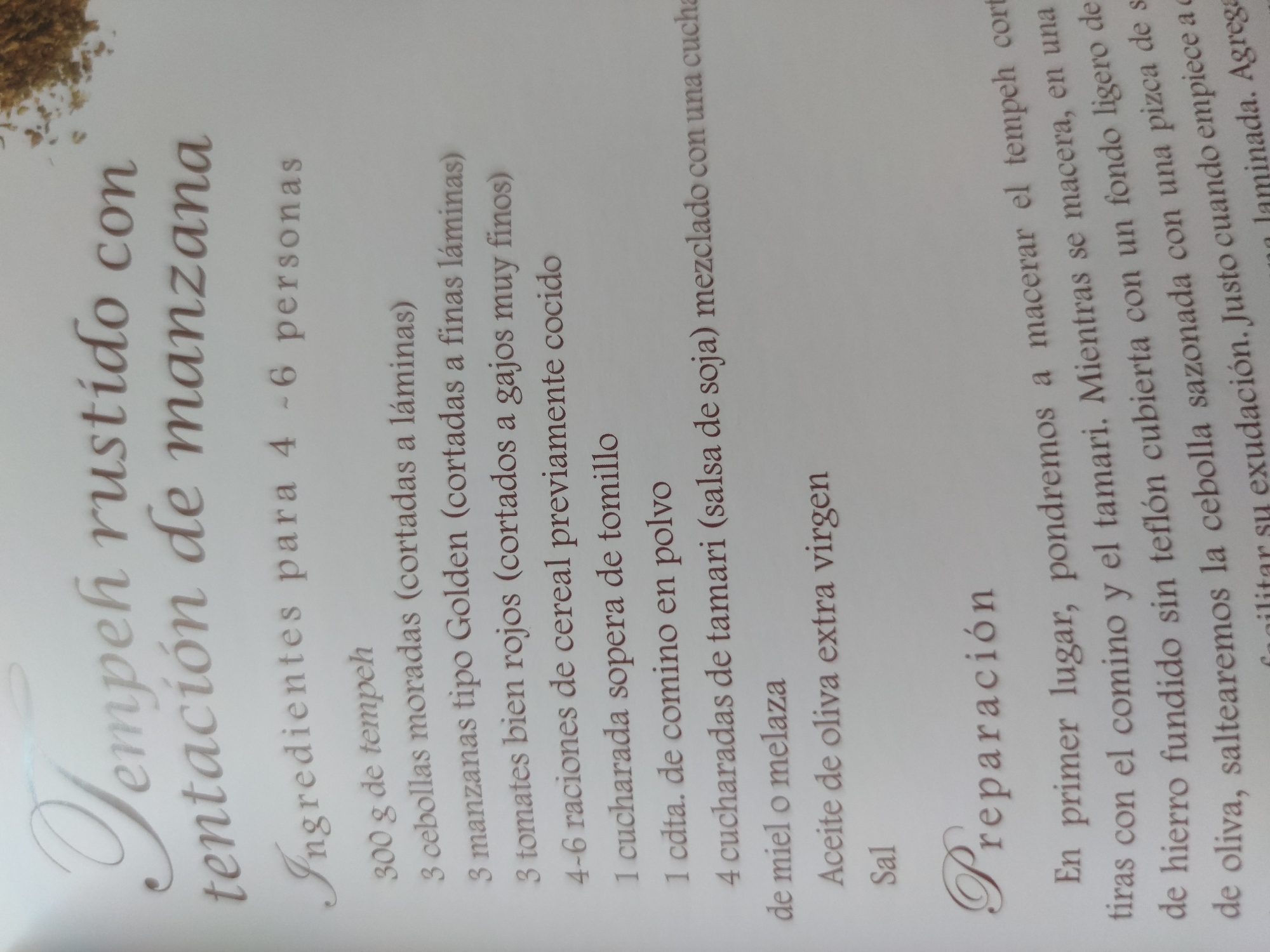 The image is a close-up shot of a page from a recipe book. The recipe is titled "Tempeh rustido con tentación de manzana," which translates to "Roasted Tempeh with Apple Temptation." Below the title is a list of ingredients for 4-6 people. The ingredients include tempeh, red onions, Golden type apples, red tomatoes, cereal rations, thyme, cumin, tamari (soy sauce), honey or molasses, olive oil, and salt. Following the ingredient list is the "Preparación" section, which provides instructions on how to prepare the dish. The instructions mention marinating the tempeh, stir-frying the onion, and seasoning with salt.