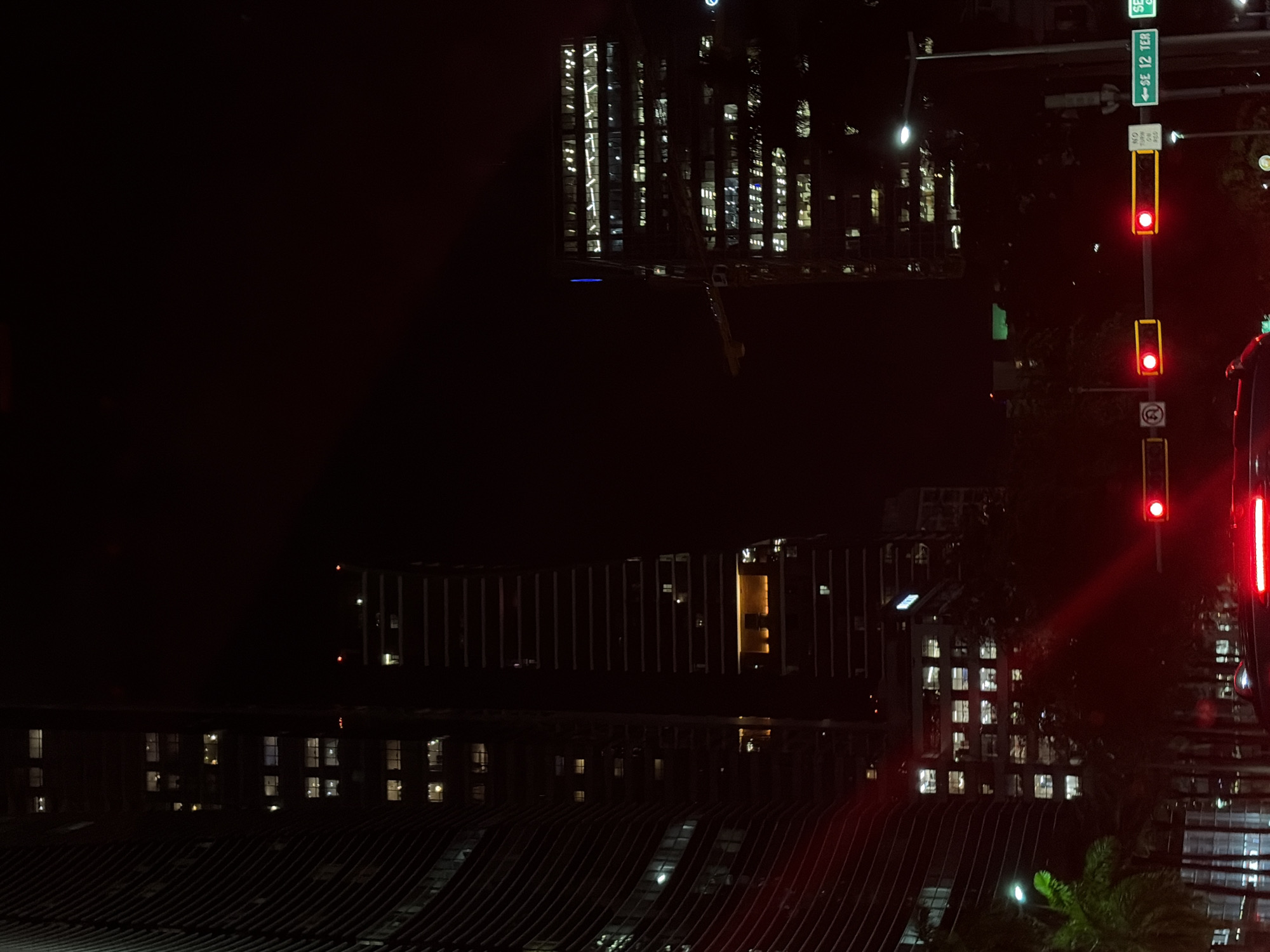 At night in Miami, USA, the image shows traffic lights with red signals on SE 12th Terrace. Buildings with lit windows are visible, including a skyscraper under construction. The area is dark, with lights from windows and street signals illuminating the scene.