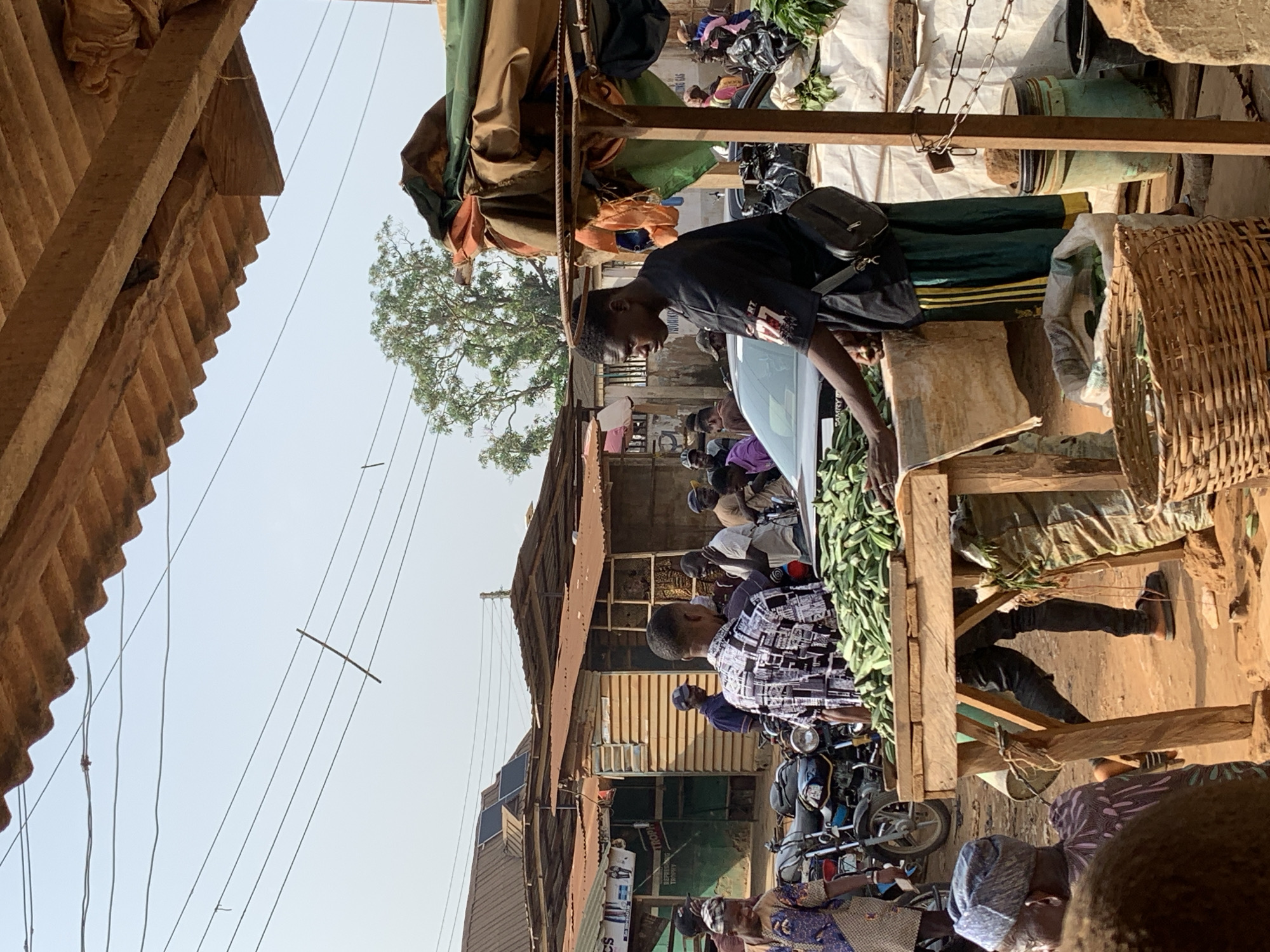 The image depicts a bustling street scene in Ikorodu, Nigeria. A young man in a black t-shirt and shoulder bag is standing near a table piled high with okra, possibly a vendor or customer at a market. Other people can be seen in the background, some wearing patterned shirts or head coverings. A street vendor in front of his table selling goods. Motorbikes are parked nearby. The environment is urban, with corrugated metal roofing and utility lines visible overhead. The scene appears to be a busy outdoor marketplace.