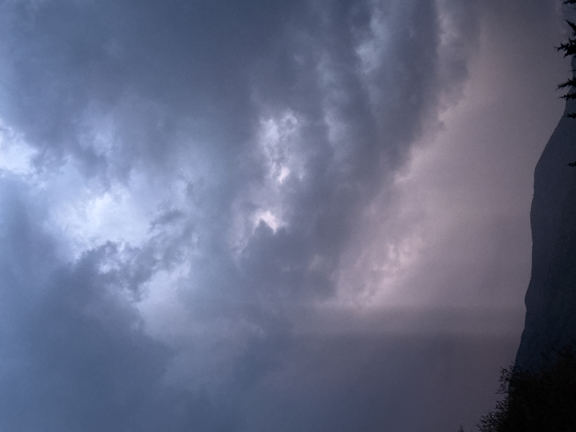 The image presents a dramatic sky dominated by dense, layered clouds, rendered in varying shades of dark blue-gray, purple, and soft pink. The upper left and central portions of the sky feature brighter, almost white, patches of cloud, indicating areas where light penetrates or reflects within the heavy cloud cover. Towards the right side, the clouds take on a distinct soft pinkish-purple hue, which could suggest a sunset or sunrise struggling to emerge through the thick atmosphere, or unique light scattering conditions.
A prominent feature on the right side of the frame is the dark, imposing silhouette of a mountain or a steep hill, its jagged profile contrasting sharply with the clouded sky. At the very bottom edge of this silhouette, the faint, textured outline of trees or other foliage can be discerned.
The scene conveys a sense of overcast weather, likely indicative of impending rain or recent stormy conditions. The time of day appears to be either early morning, late evening, or a deeply overcast midday, due to the subdued lighting and specific color palette. The presence of a mountain silhouette aligns with the provided location context of Benasque, Spain, a town situated in the mountainous Pyrenees region. No specific human activity or visible text is present in the image.