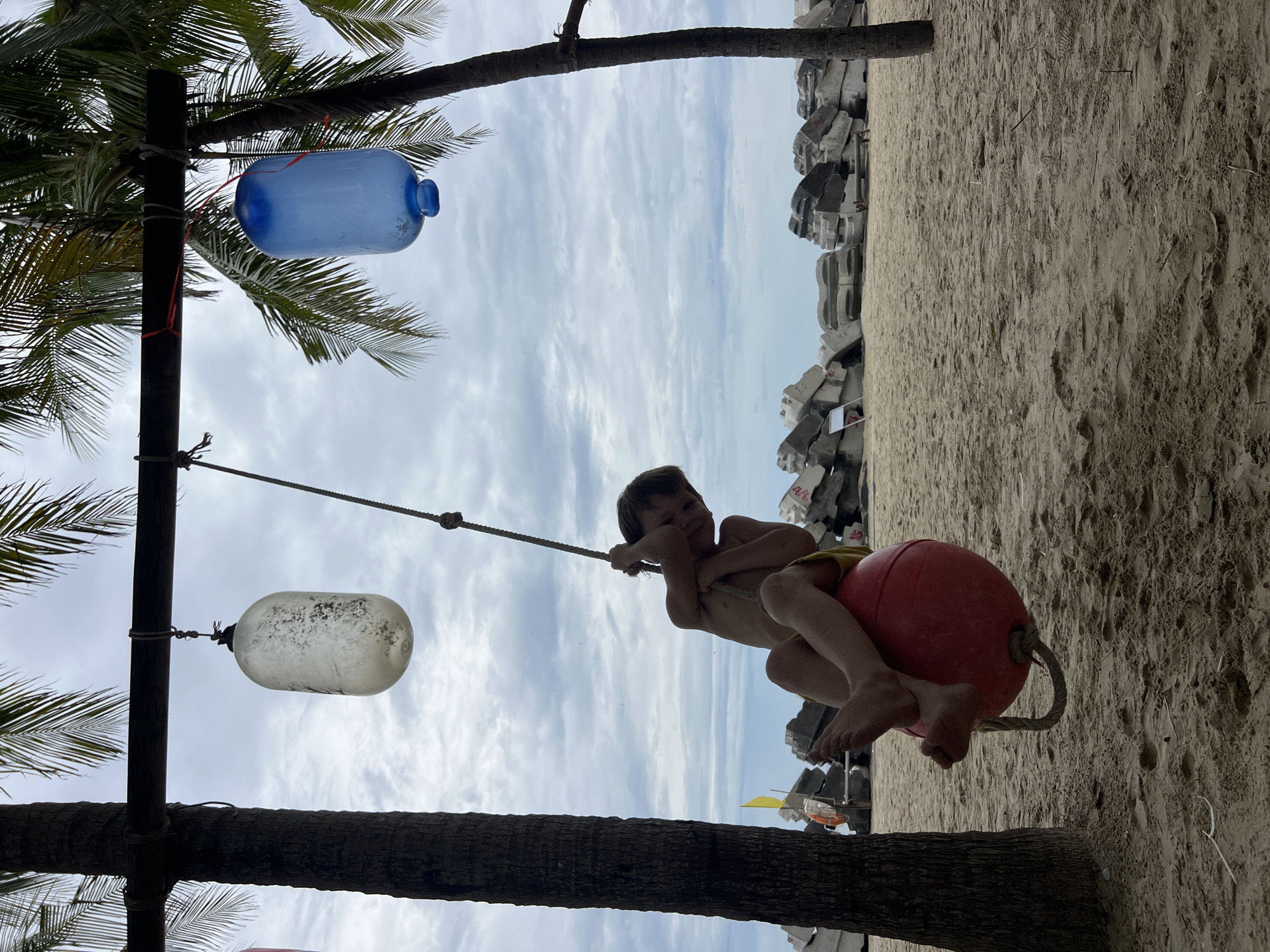 A young boy is swinging on a red ball swing suspended from a wooden beam between two palm trees on a sandy beach. He is wearing yellow shorts and is holding onto the rope of the swing, with his legs dangling. In the background, there is a cloudy sky, the ocean, and a line of large concrete blocks. Two large, translucent containers, one white and one blue, are hanging from the same wooden beam above the swing. The scene appears to be on a beach, possibly near a protected area of the coast, given the presence of the concrete blocks.