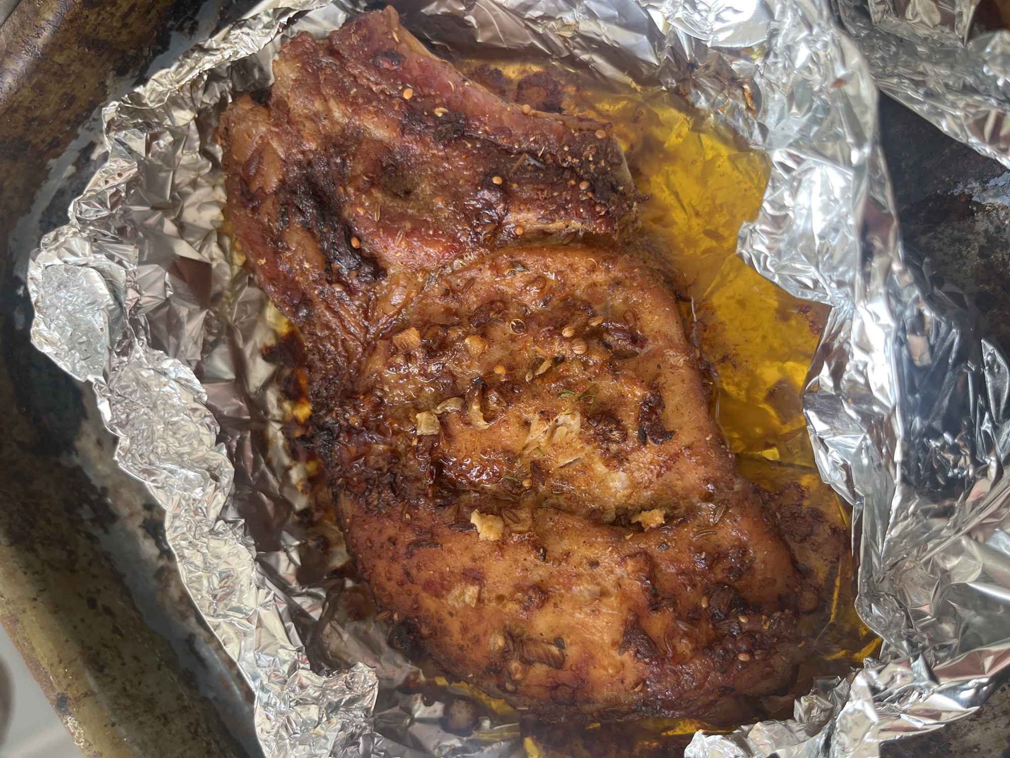 This is a close-up, overhead shot of a single, cooked pork chop resting on crumpled aluminum foil within a dark, possibly cast-iron, baking pan. The pork chop is golden brown with darker, seared edges and appears to be seasoned with herbs and spices, some of which are visible as small flecks on the surface. A pool of clear, yellow liquid, likely rendered fat or juices, surrounds the bottom portion of the chop. The pan shows signs of wear and some residue, suggesting it has been used for cooking. The background is out of focus, but hints at a kitchen environment. There are no people or animals visible in the image. The scene does not depict any specific location in the city of Chelyabinsk, Russia.