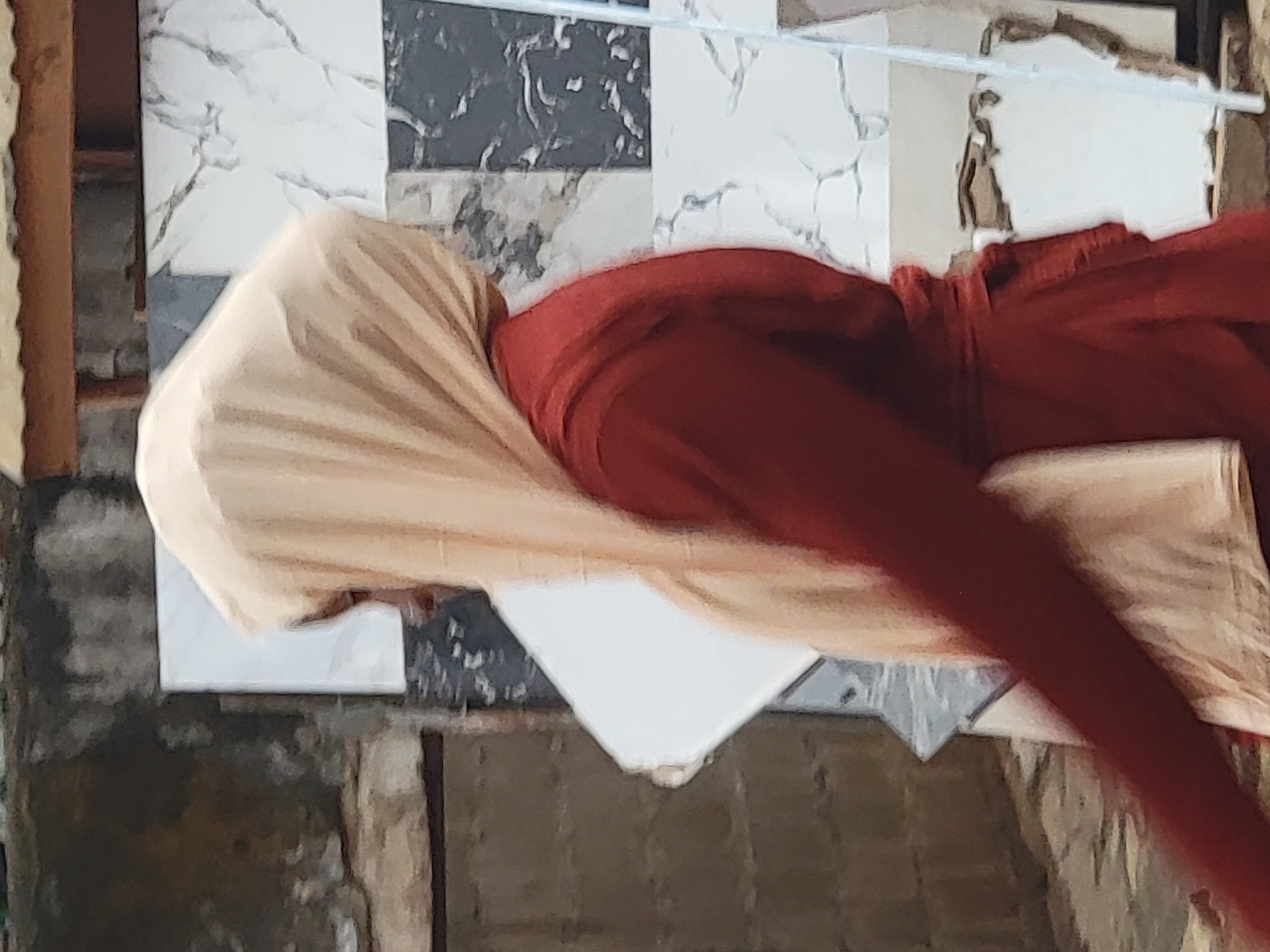 This image captures a person draped in flowing fabrics, positioned vertically against a tiled wall. The individual's head is covered by a light beige fabric, which cascades down, partially obscuring the upper body. The rest of the visible body is covered in a rich red fabric, possibly trousers or a full garment, which extends downwards and slightly to the right. The person's face is not visible.
The background primarily consists of a wall adorned with an assortment of tiles. These include white tiles with prominent grey/black marble-like veining, as well as some solid black/grey tiles with similar subtle patterns, and a plain white rectangular tile in the lower-middle section. A thin, white horizontal line, possibly a wire or clothesline, is visible across the upper portion of the background. To the far left, a blurred, dark brown, textured vertical element, possibly a wooden beam or part of a structure, can be seen. The foreground and other peripheral parts of the scene are blurred, revealing earthy tones and indistinct shapes that suggest ground, other walls, or rustic structures.
The setting appears to be an outdoor, possibly residential or communal, area. The lighting suggests it is daytime. There is no explicit activity taking place beyond the person's pose. No emotions are discernible due to the obscured face, and no text is visible within the frame. The overall scene conveys a candid moment in an everyday environment in Maiburiji, Nigeria.