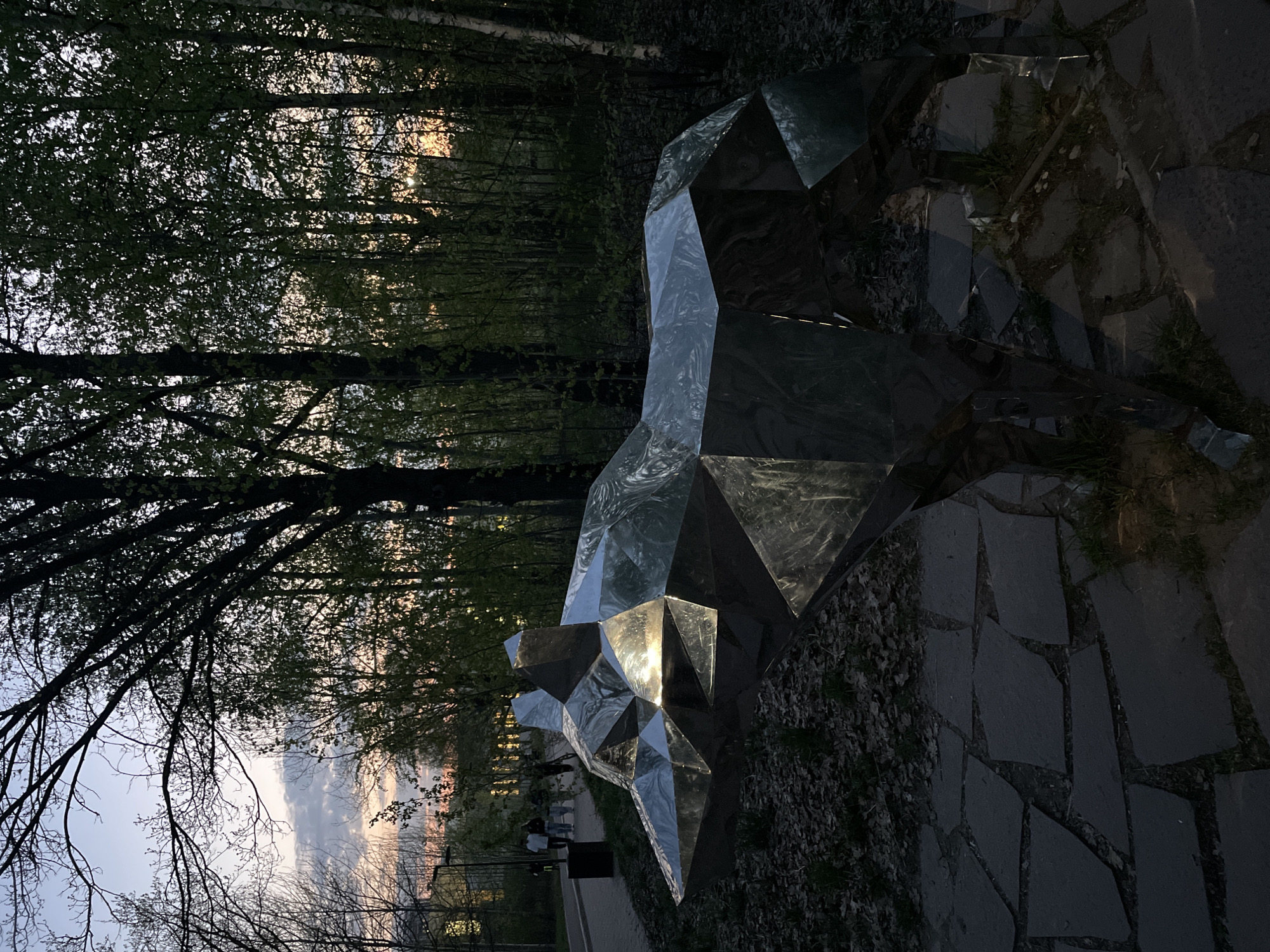 The image depicts a polygonal metal sculpture resembling a wolf or bear head, positioned on a paved area with grass and foliage. The sculpture reflects sunlight, creating bright highlights on its facets. In the background, there is a dense wooded area with trees and hints of sunset. The sky appears to be a pale blue with traces of pink and orange near the horizon. In the lower left, there is a glimpse of a walkway with a few people and a street lamp, suggesting a park or urban setting. The location is Kazan, Russia.