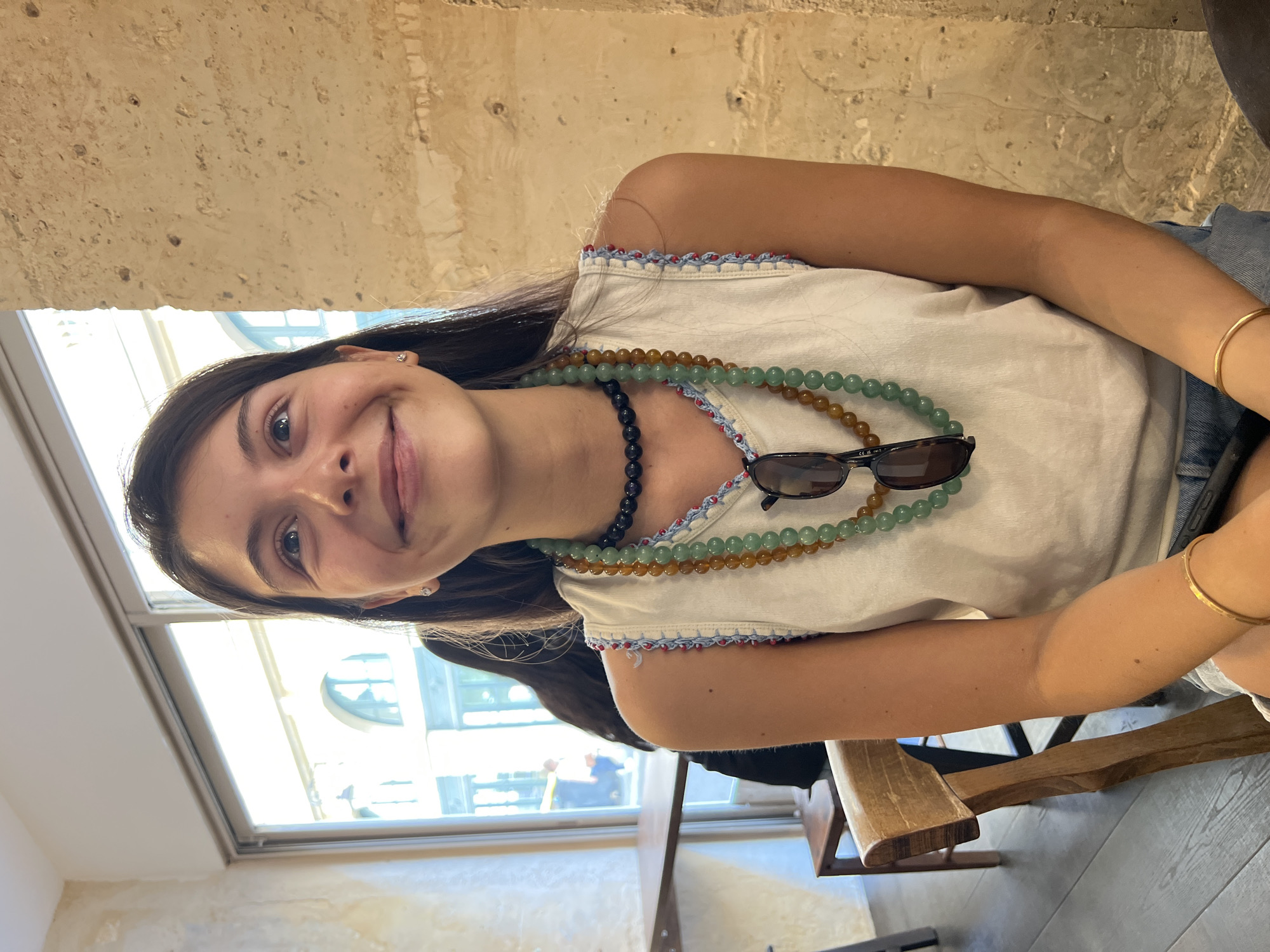 This is a portrait-oriented media file of a young woman with dark hair, looking at the camera with a slight smile. She is wearing a white top with decorative trim and several beaded necklaces. Sunglasses are perched on her shirt. She has a gold bracelet on her right wrist. The background appears to be an indoor setting with a textured wall and a window showing a bright, possibly daytime scene. The overall lighting suggests it's not nighttime. There is no visible text in the image.
