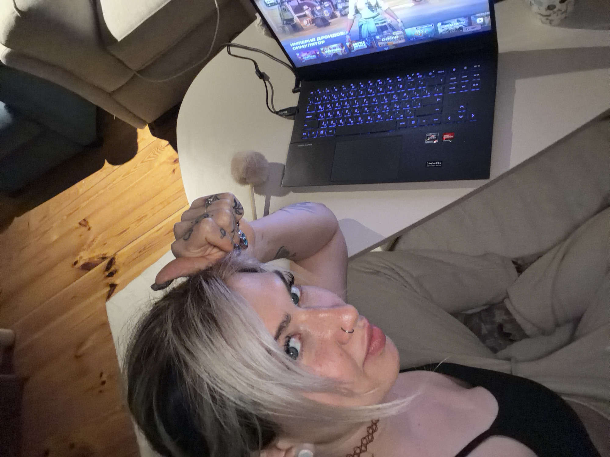 A woman with dyed blonde and black hair is lying on a light-colored sofa, holding her hand to her head, with a laptop in front of her on a white table, the screen displaying a video game. The keyboard of the laptop is backlit with blue light. The background includes a wooden floor and a glimpse of another chair. The image appears to be taken indoors. There is no indication of the location being in Budva, Montenegro.