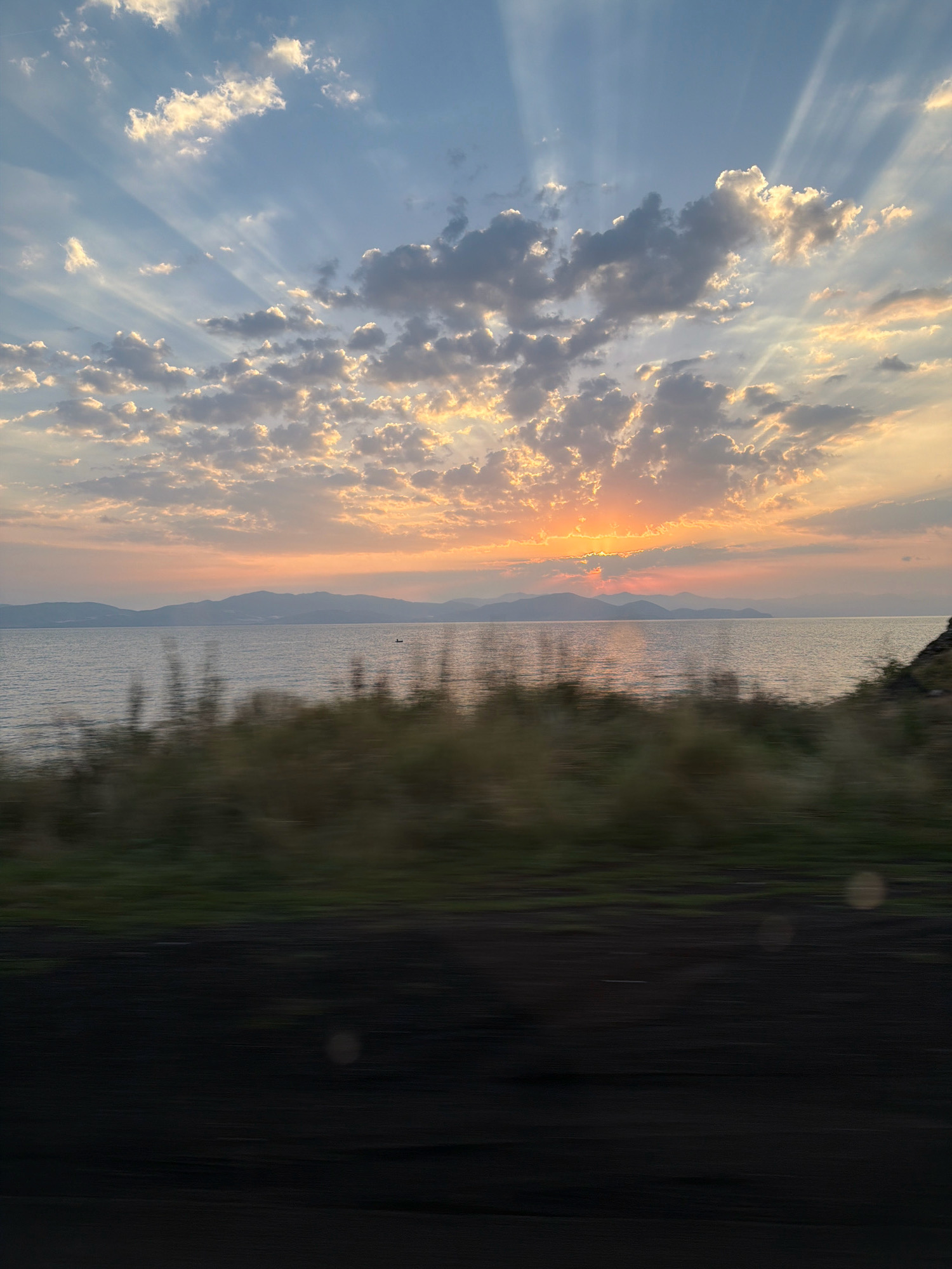 The image captures a scenic sunset over a body of water, likely Lake Sevan, near Berdkunk, Armenia. The sky is a canvas of soft blues and oranges, with rays of sunlight piercing through scattered clouds, creating a dramatic crepuscular effect. A distant mountain range forms the horizon, its silhouette softened by the twilight. The water is calm, reflecting the warm hues of the sky. In the foreground, blurred vegetation suggests movement, perhaps from a vehicle passing by, as indicated by the horizontal motion blur. A small boat is visible on the water, adding a subtle human element to the vast landscape. The overall impression is one of tranquility and natural beauty at the end of the day.