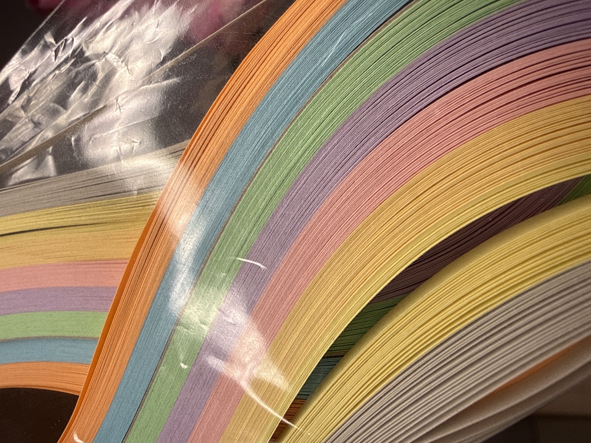 A close-up captures a collection of thin strips of paper in various pastel colors, including orange, blue, green, purple, pink, yellow, and gray. These strips are arranged together, creating a layered, arched pattern. The top portion of the image shows the collection wrapped in clear plastic. The location is not discernible from the image.