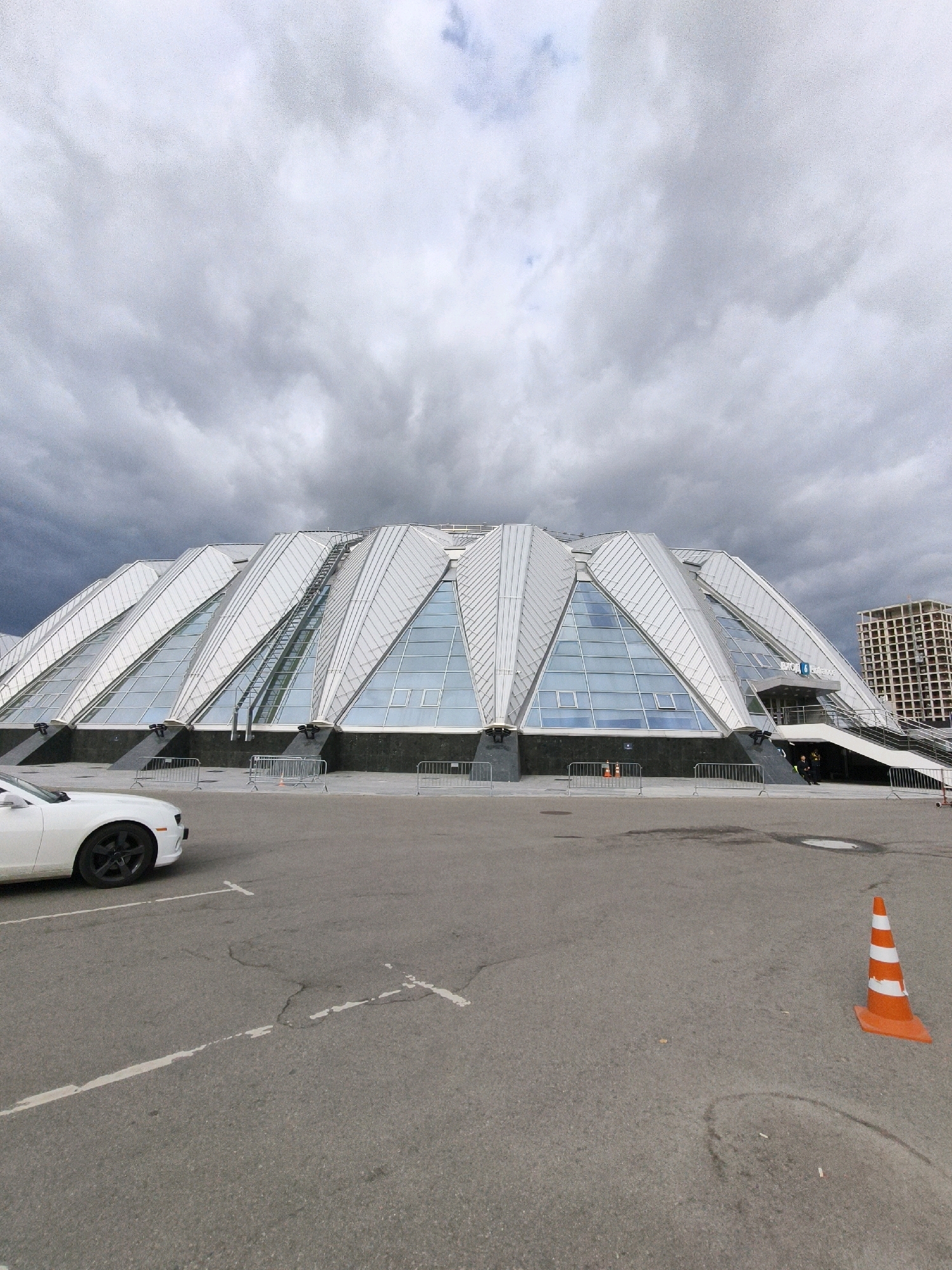 The image captures the exterior of the Ice Palace in Moscow, Russia, under a heavily overcast sky. The Ice Palace is a large, modern building with a distinctive faceted roof structure made of white panels and triangular glass windows. In the foreground, a white car, identified as a Chevrolet Camaro, is parked on an asphalt lot marked with white parking lines. An orange traffic cone with white stripes stands to the right of the car. In the background, a tall, unfinished building with exposed concrete and rebar can be seen. Several metal barriers are placed in front of the Ice Palace, and a few individuals are visible near the entrance stairs. The overall impression is a somewhat stark and utilitarian scene, likely on a cloudy day.